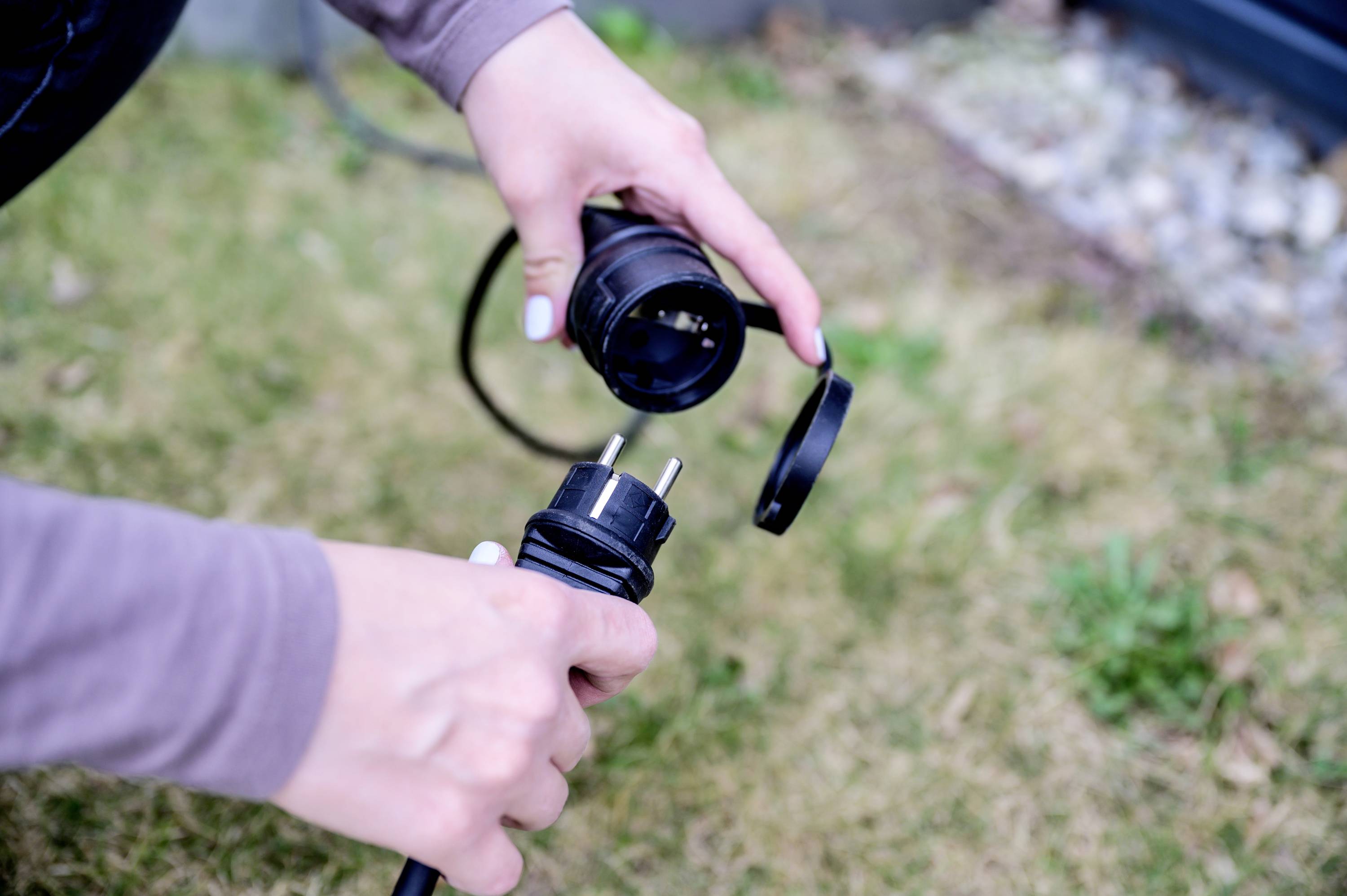 Eine Person hält einen Stecker und eine Steckdose in der Hand, bereit zum Verbinden. Der Hintergrund zeigt einen Rasenbereich.