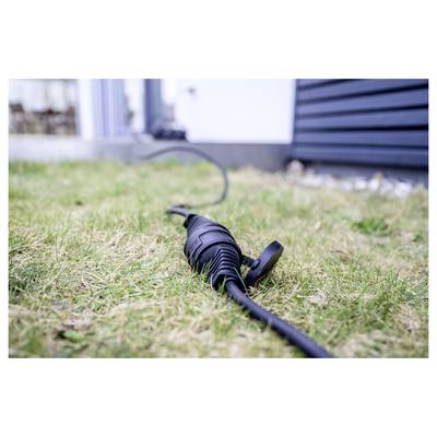 Nahaufnahme eines schwarzen Außenelektrokabels mit schützender Steckerdose, das auf trockenem Gras neben einer Hauswand und Verkleidung liegt.