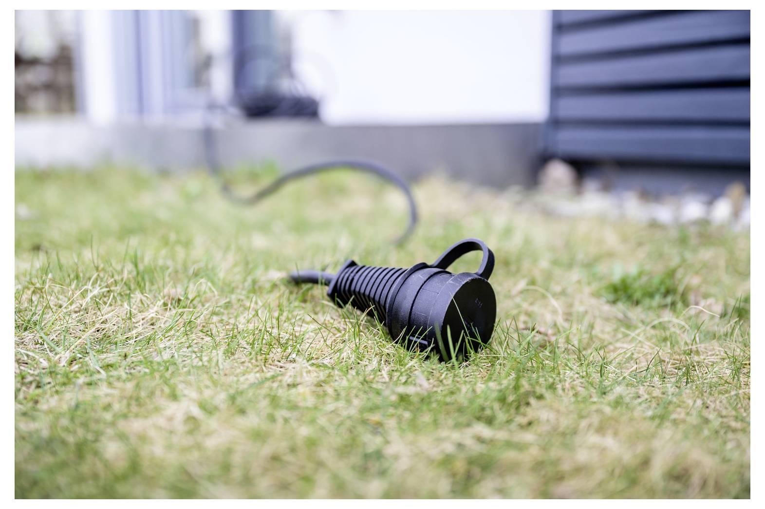 Schwarzes Stromkabel mit Stecker liegt auf Gras im Freien, mit einem verschwommenen Gebäude im Hintergrund.