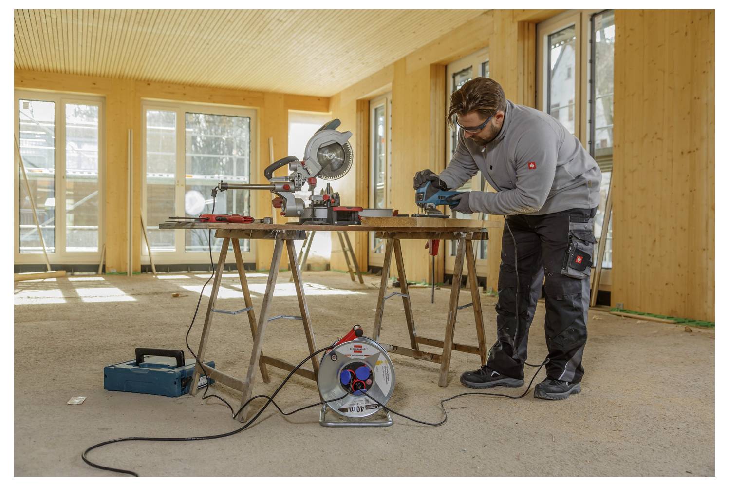 Ein Zimmermann, der eine Schutzbrille trägt, arbeitet mit einem Elektrowerkzeug an einer Werkbank in einem teilweise fertiggestellten Holzzimmer.