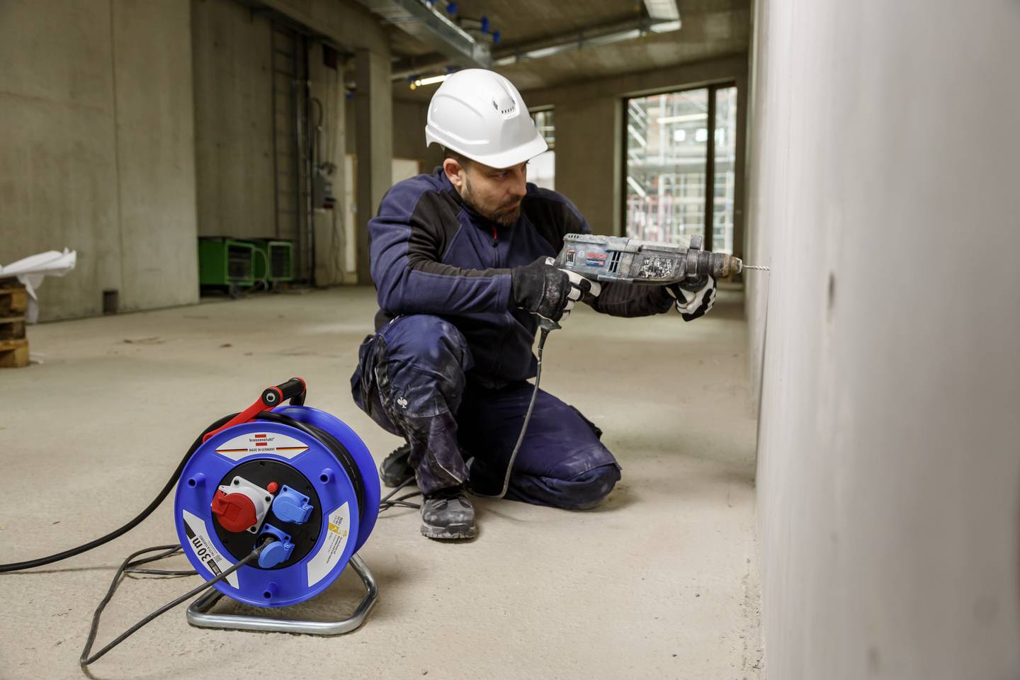 Ein Arbeiter in Schutzkleidung und Helm bohrt ein Loch in eine Betonwand auf einer Baustelle. Neben ihm steht eine Kabeltrommel.