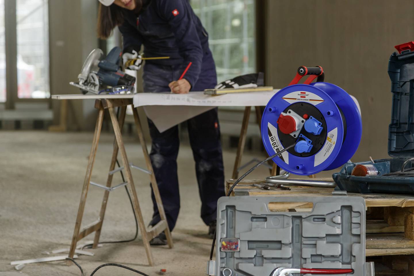 'Person arbeitet konzentriert an einem Tisch auf einer Baustelle, mit Bauzeichnung und Kreissäge. Im Vordergrund liegt eine Kabeltrommel.'