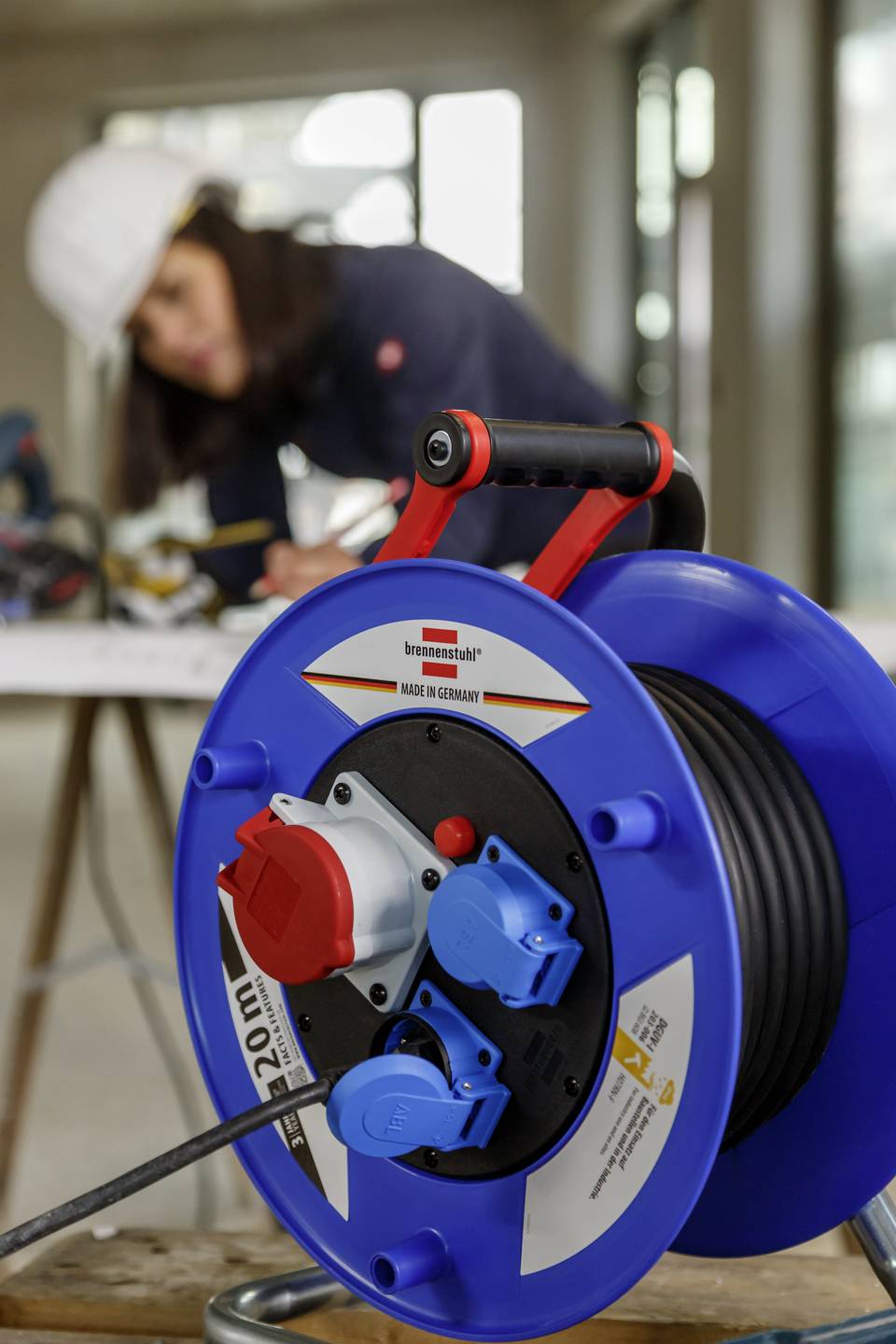 Eine Frau im Bauhelm arbeitet im Hintergrund an einem Tisch. Im Vordergrund steht eine blaue Kabeltrommel.