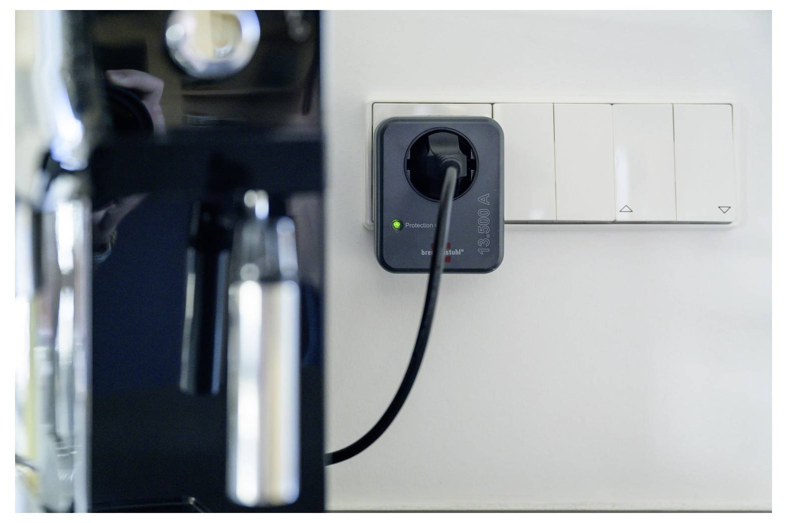 Eine eingesteckte Kaffeemaschine in der Nähe einer Wandsteckdose mit einem leuchtenden Steckeradapter und weißer Schaltertafel an einer Küchenwand.