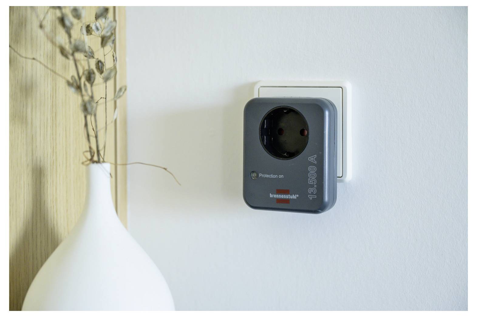 Eine an der Wand montierte elektrische Steckdose mit einem eingesteckten Adapter auf einer weißen Wand, neben einer weißen Vase mit getrockneten Stielen.