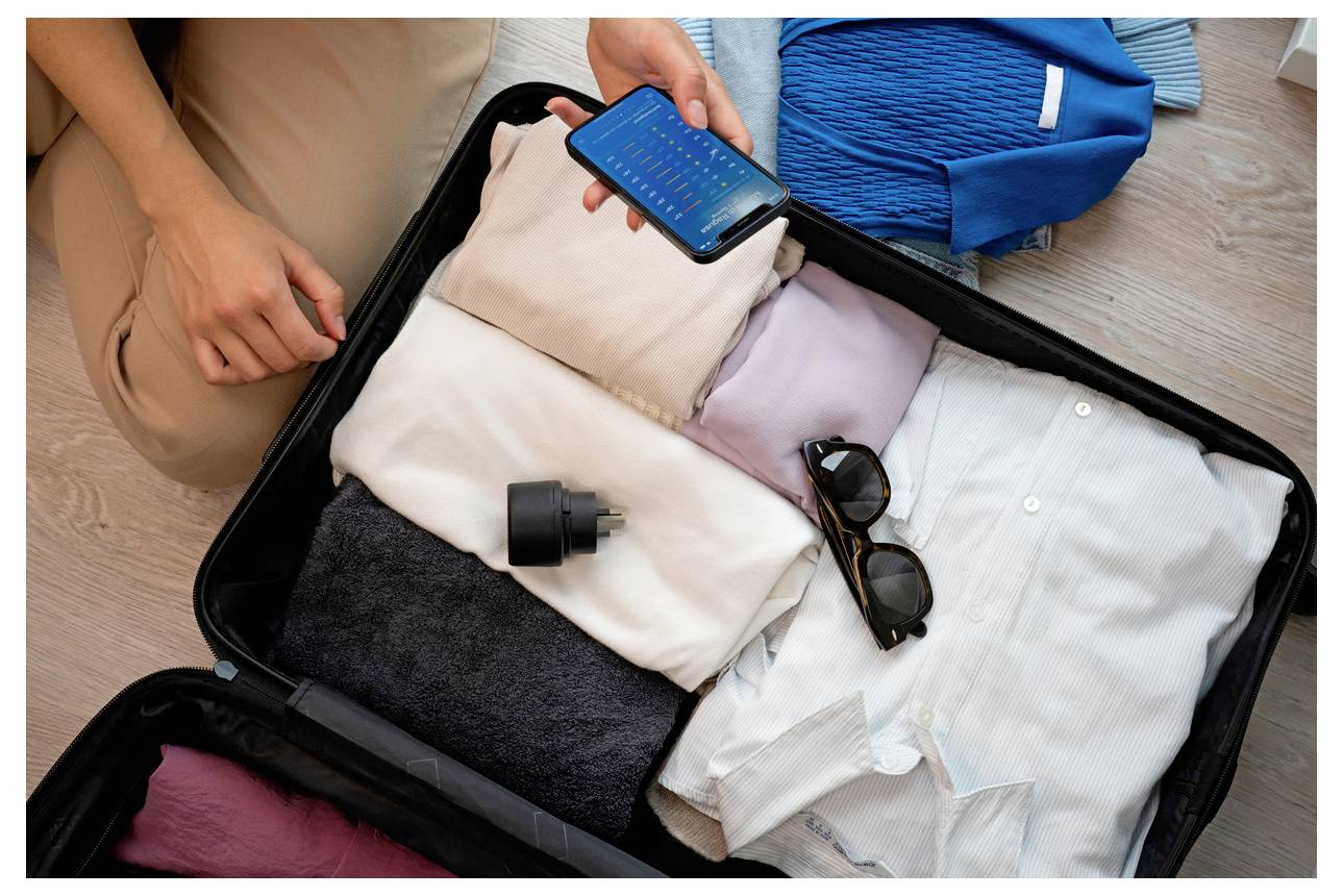 Person packt einen offenen Koffer mit gefalteter Kleidung, Sonnenbrille und einem Telefon und bereitet sich auf eine Reise vor.