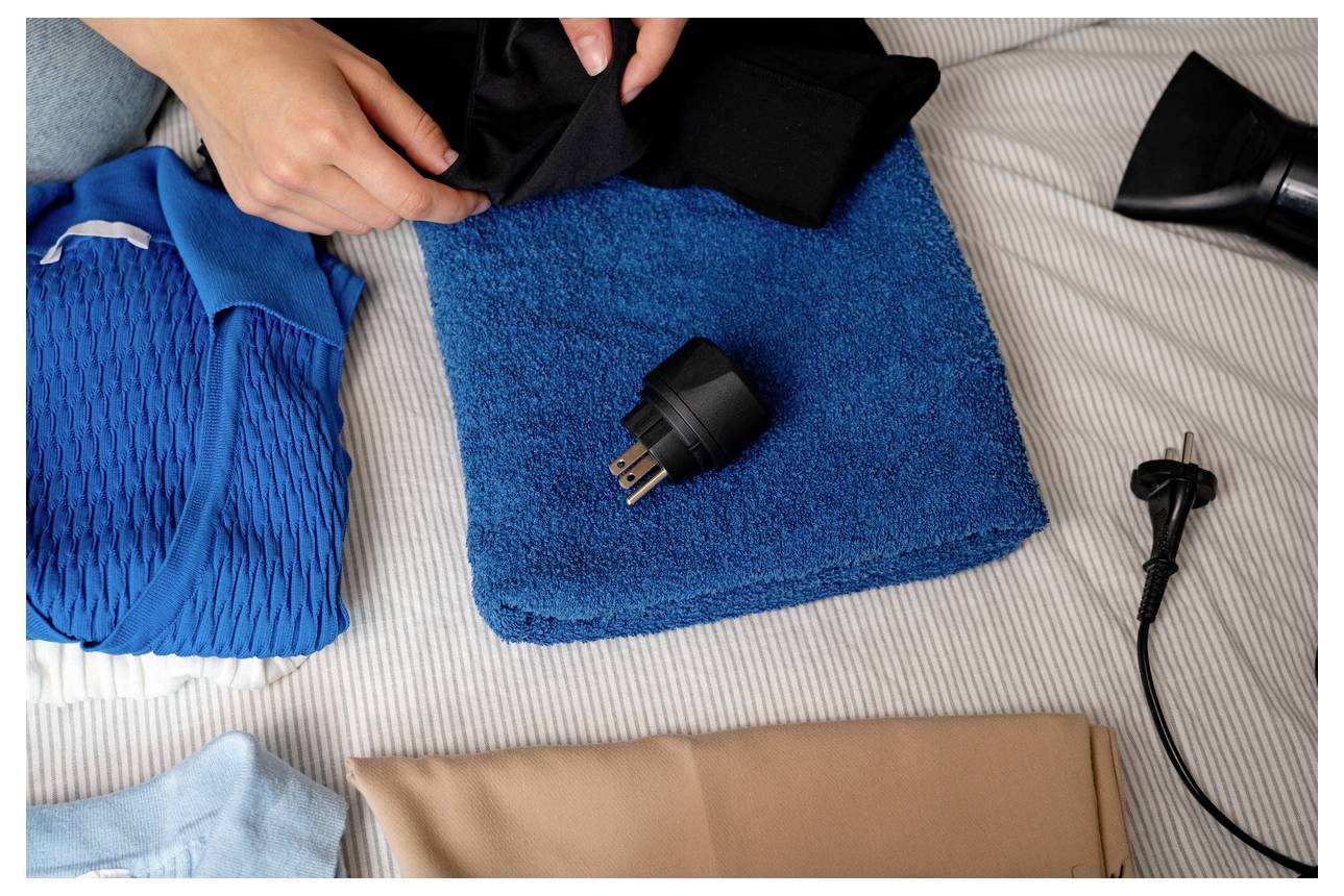 Person, die Kleidung und ein schwarzes Stromkabel auf einem Bett packt, was auf das Packen für eine Reise mit einem Elektrostecker oder Ladegerät hindeutet.