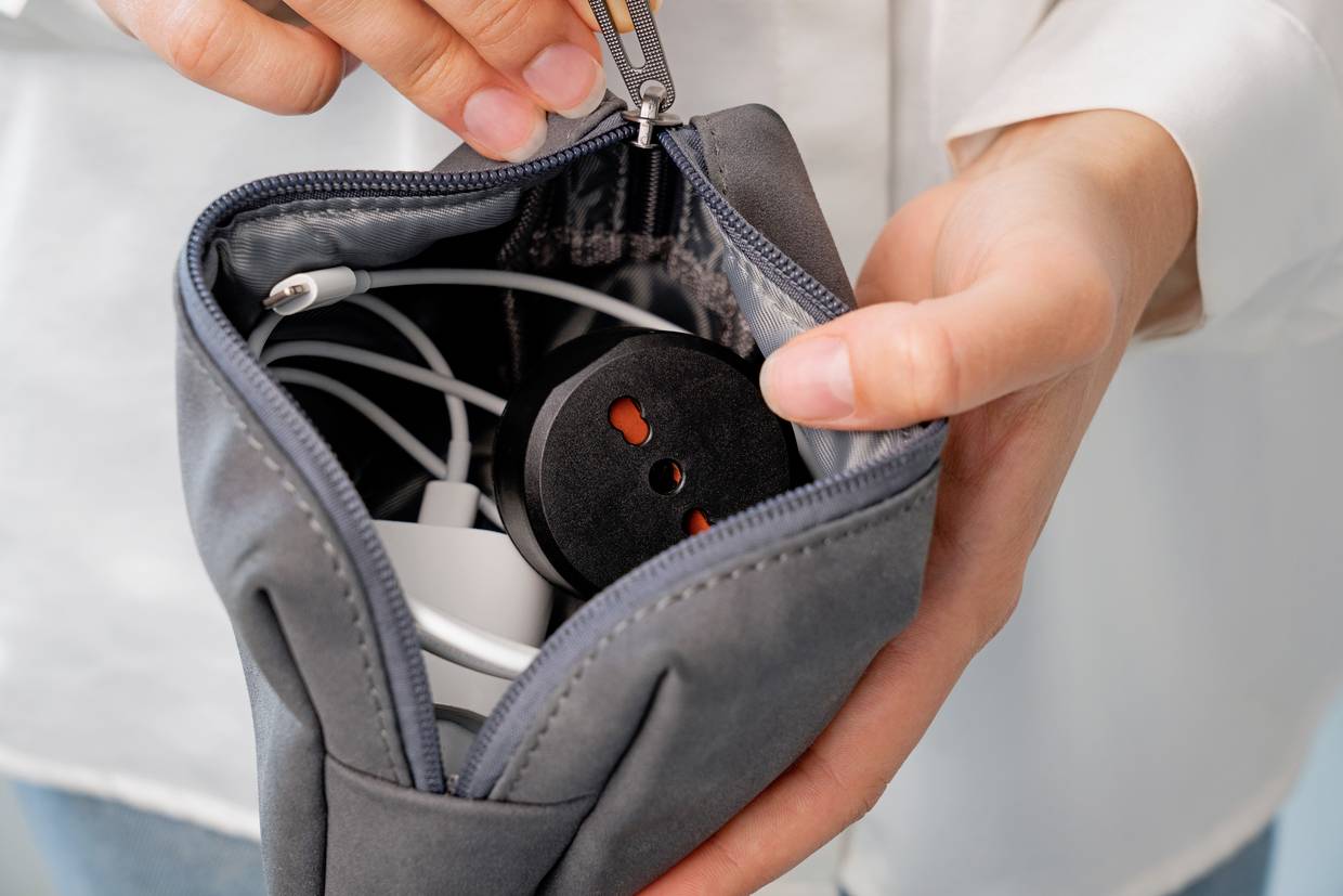 Person öffnet eine graue Tasche, die Kabel und einen Adapter enthält. Hände halten den Reißverschluss. Nahe Aufnahme.