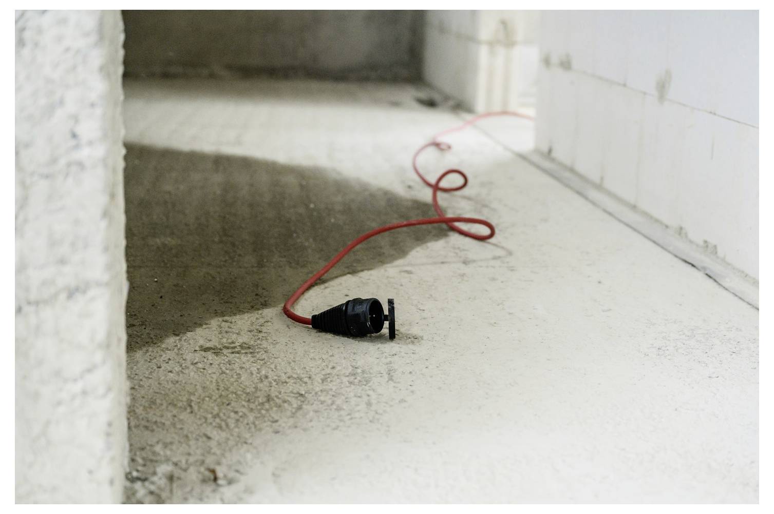 Ein rotes Verlängerungskabel mit einem schwarzen Stecker liegt auf einem staubigen Betonboden in einem unfertigen Zimmer.