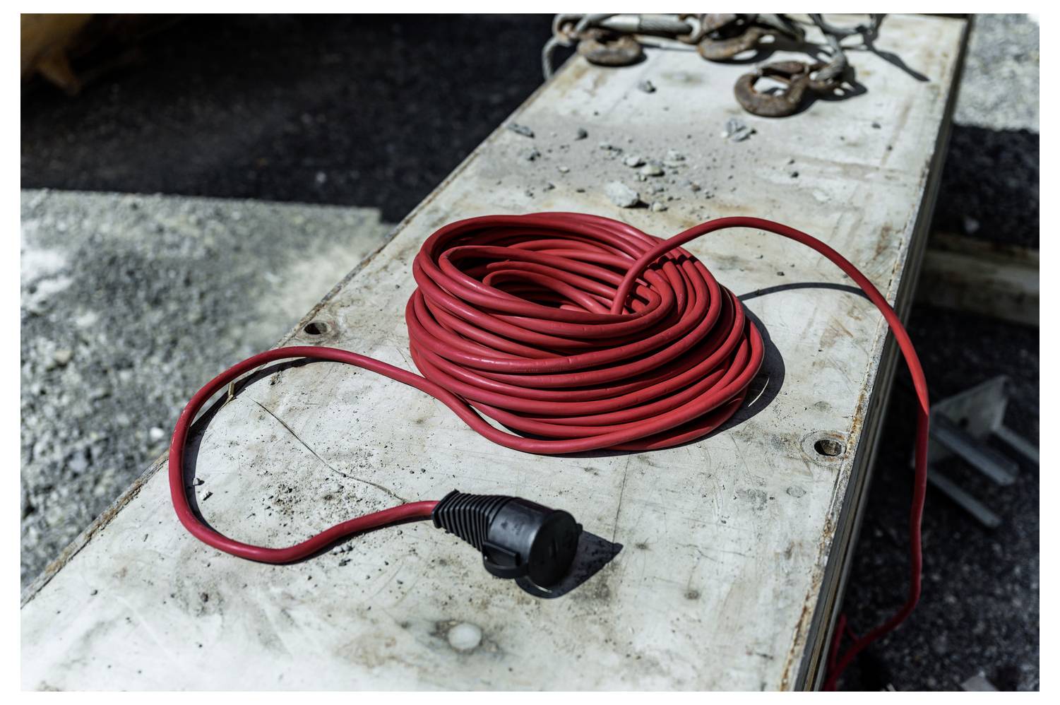 Aufgerolltes rotes Stromkabel mit schwarzem Stecker, das auf einer verwitterten Betonoberfläche im Freien liegt.