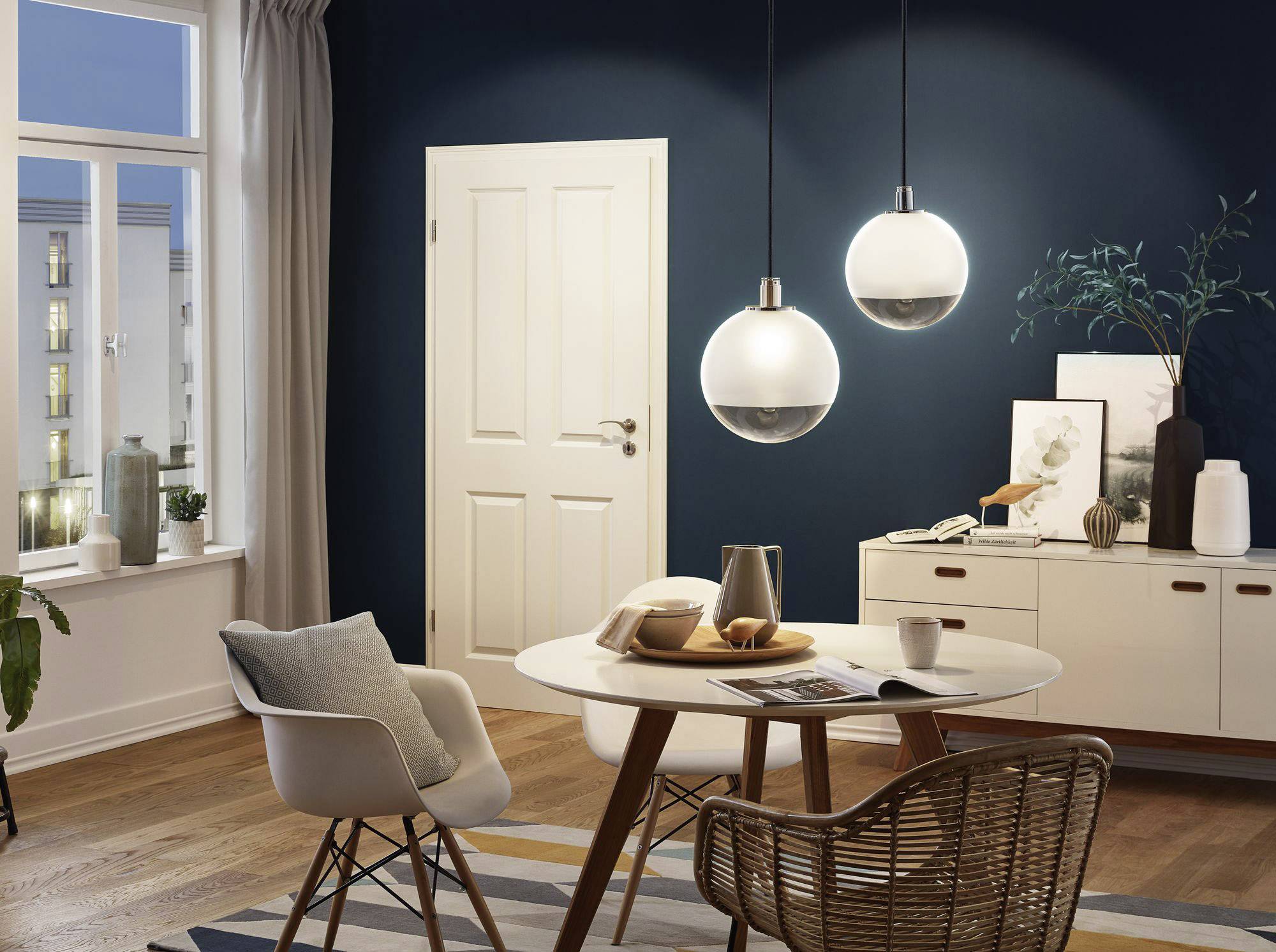 Esszimmer mit rundem Tisch, vier Stühlen und moderner Beleuchtung. Dunkle Wand, Fenster mit Vorhängen, hölzerner Boden und Kommode im Hintergrund.