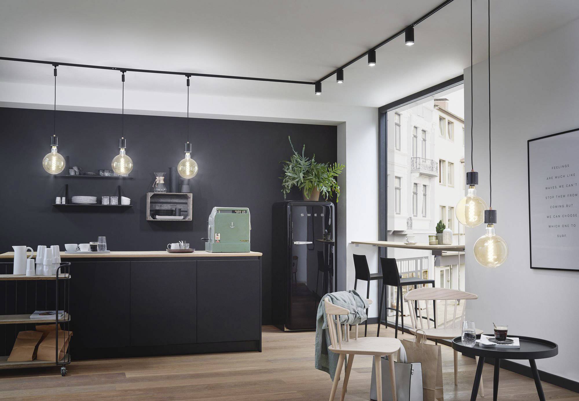 Moderne Küche mit schwarzer Wand und Arbeitsfläche. Hängelampen beleuchten den Raum, ein grüner Kühlschrank steht links, Pflanzen im Fenster.