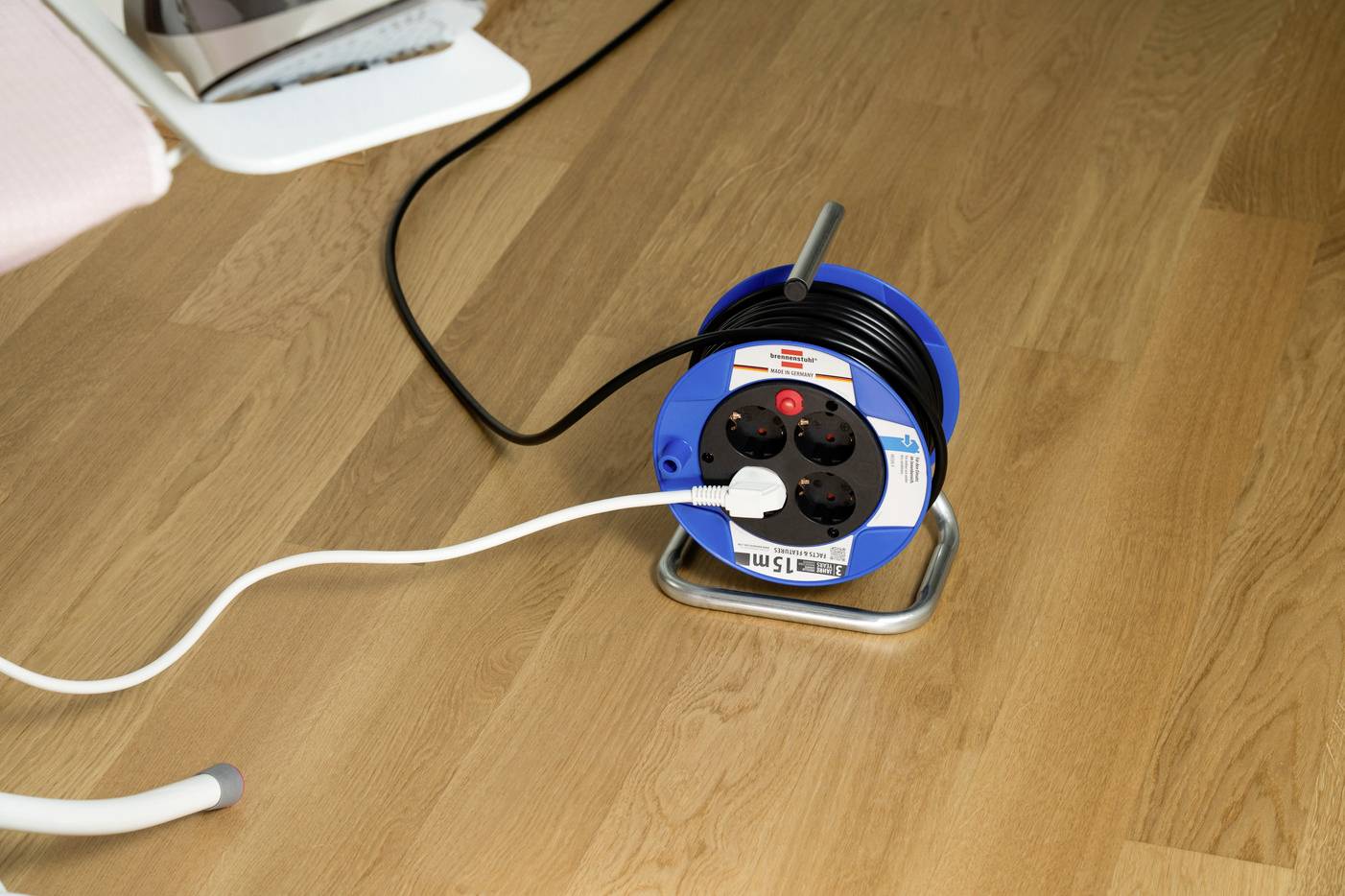 Das Bild zeigt eine blaue Kabeltrommel auf einem Holzboden. Ein weißes und ein schwarzes Kabel sind in die Steckdosen der Trommel eingesteckt.