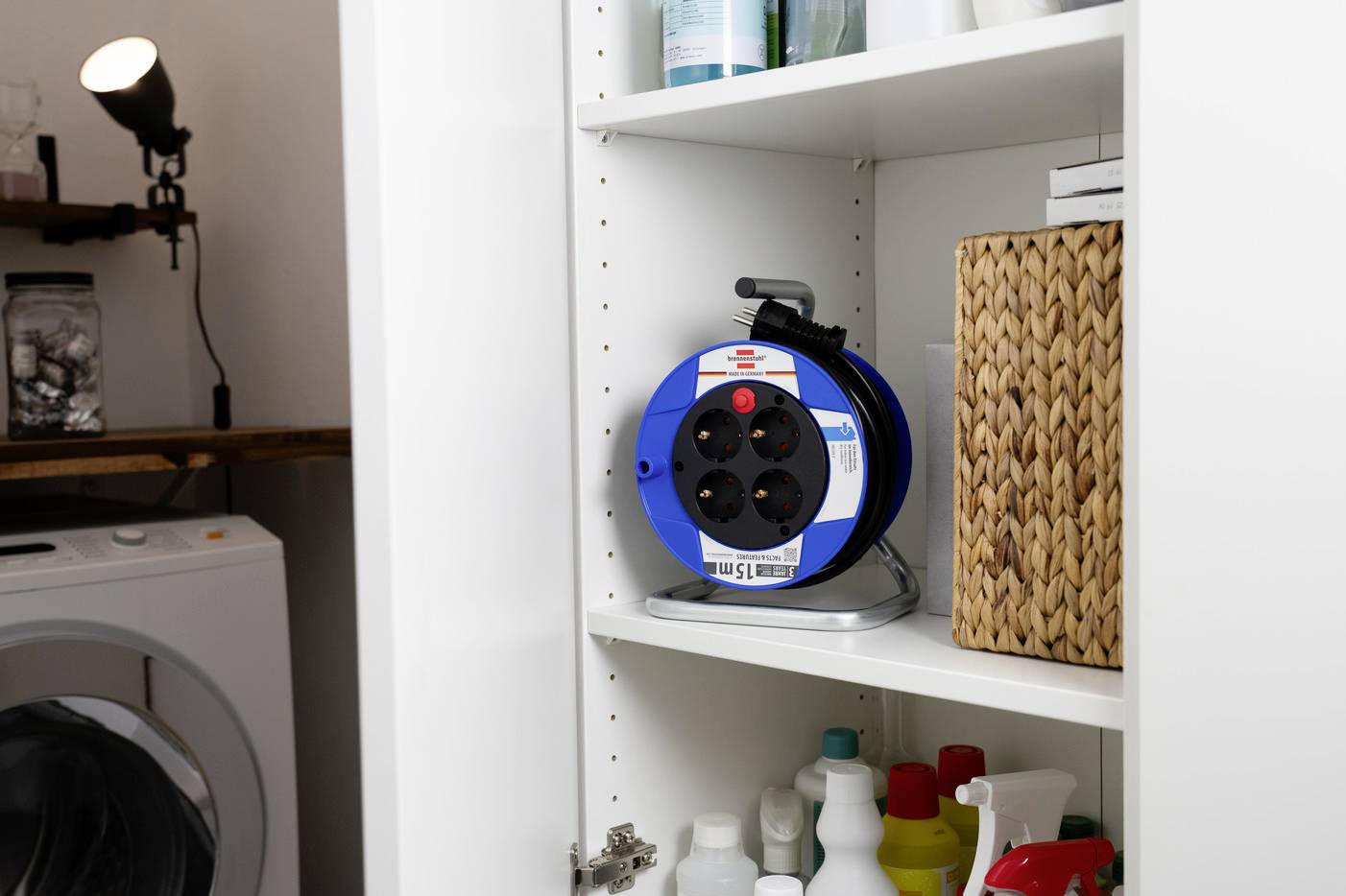 Ein geöffneter Schrank zeigt Reinigungsmittel und eine Kabeltrommel. Im Hintergrund ist eine Waschmaschine sichtbar.