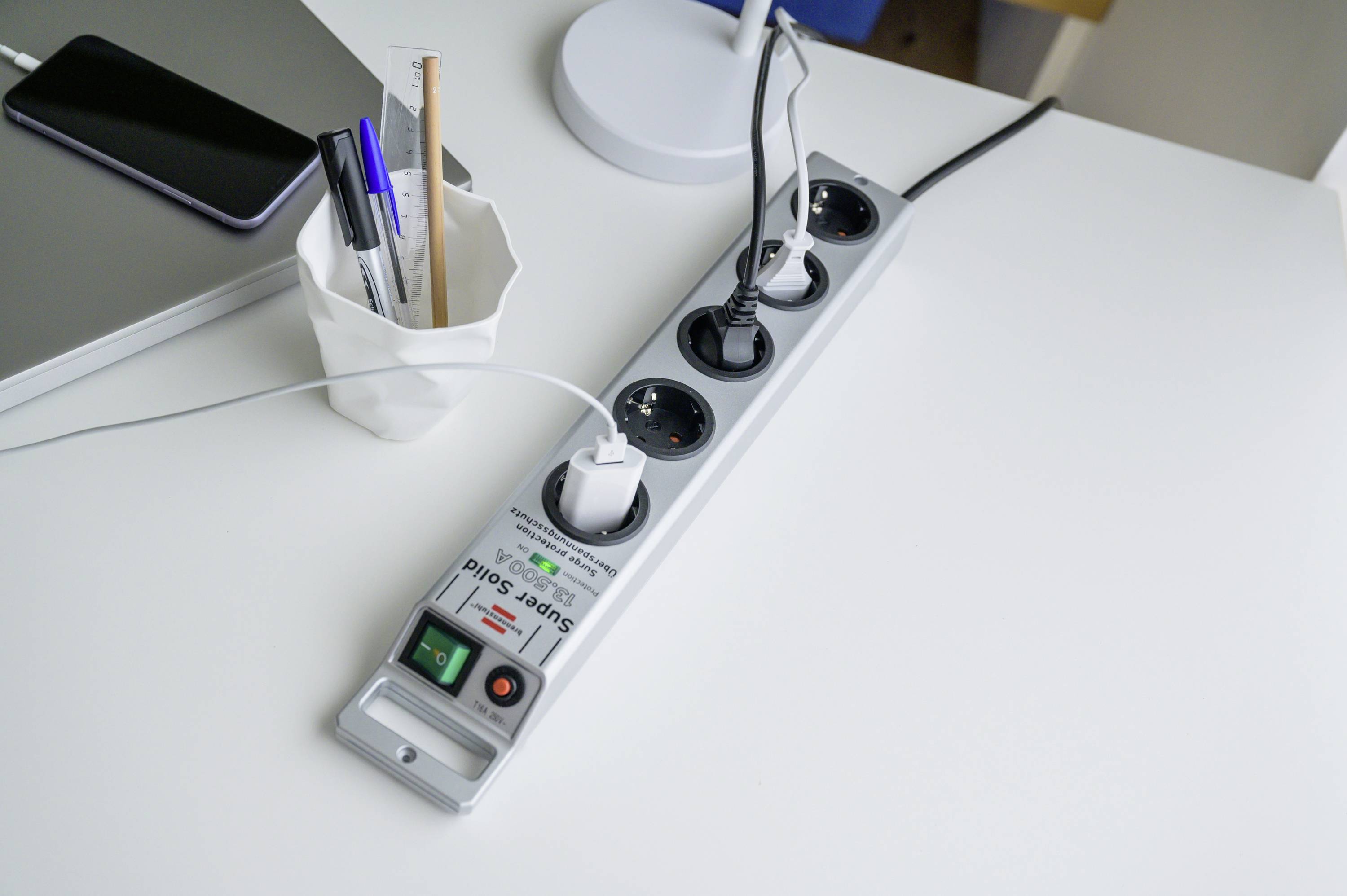 'Verlängerungskabel mit mehreren Steckdosen auf einem Schreibtisch, mit verschiedenen eingesteckten Ladekabeln und daneben liegenden Büromaterialien.'