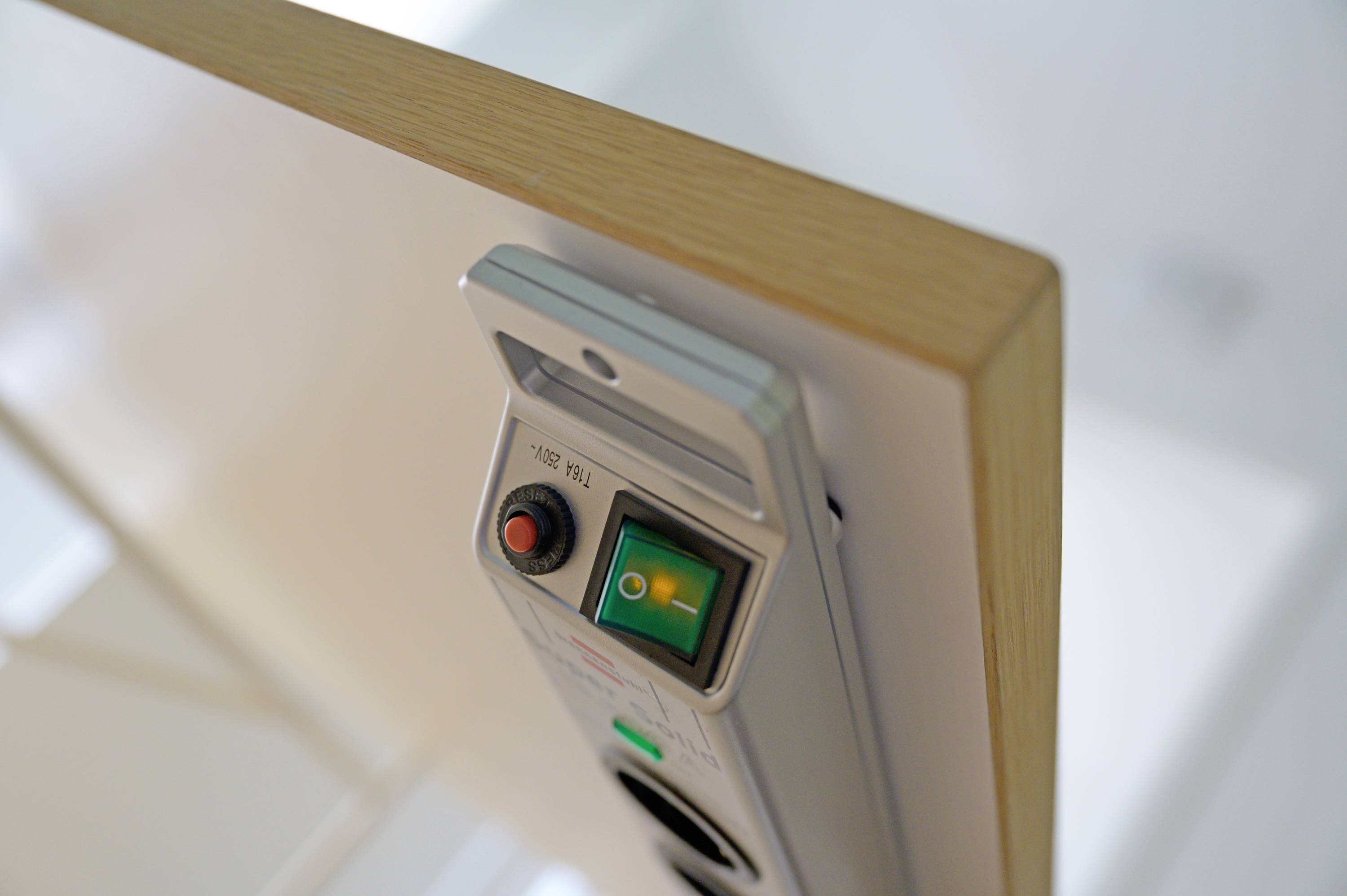 Ein Bedienfeld an einer Tischkante zeigt einen roten Knopf und ein grünes Lämpchen an einer glatten Oberfläche, möglicherweise zur Steuerung eines Geräts.