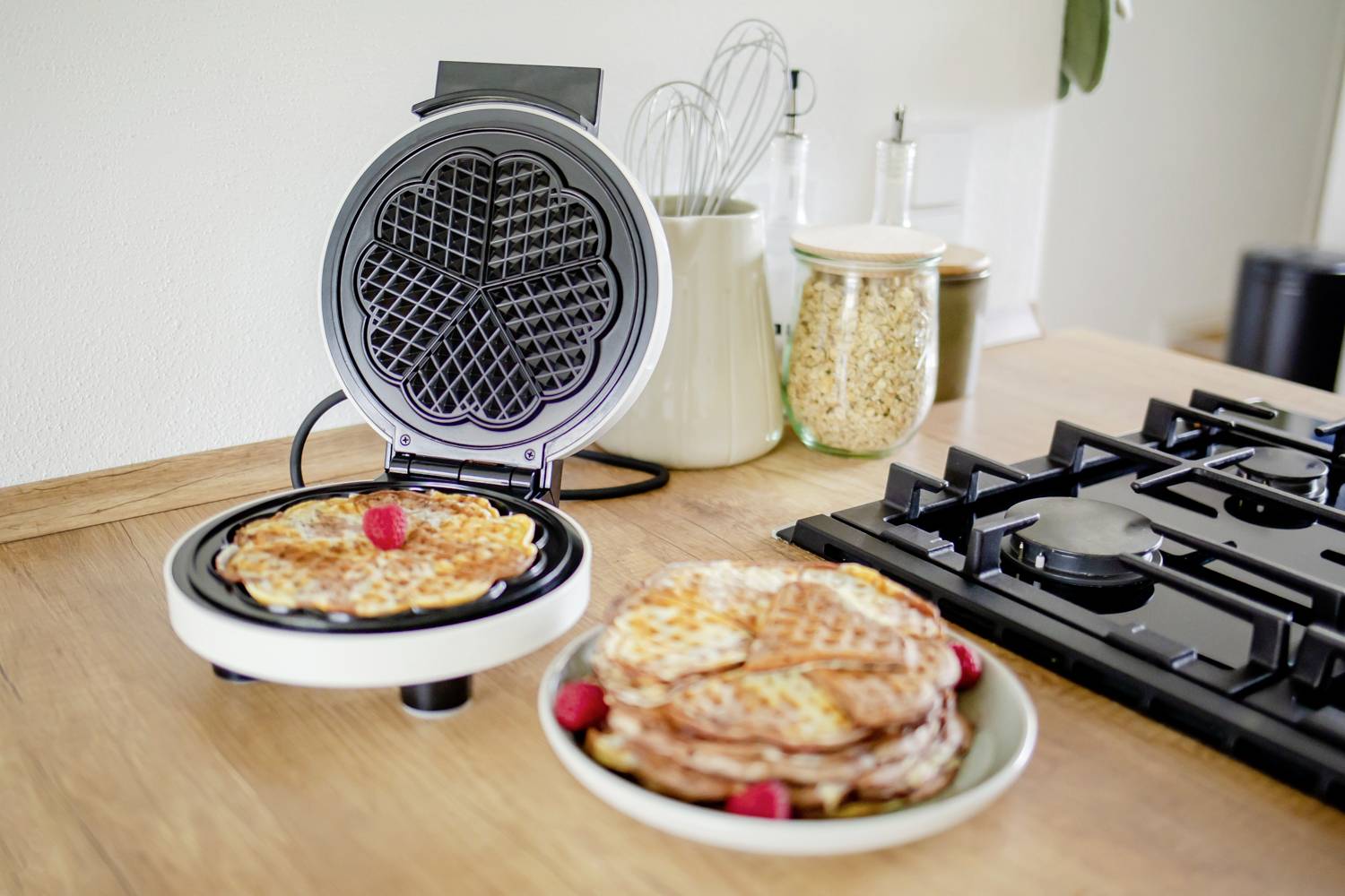 Waffeleisen mit geöffneter Klappe auf einer Holzarbeitsfläche. Eine fertige Waffel im Gerät und ein Teller mit Waffeln und Himbeeren daneben.