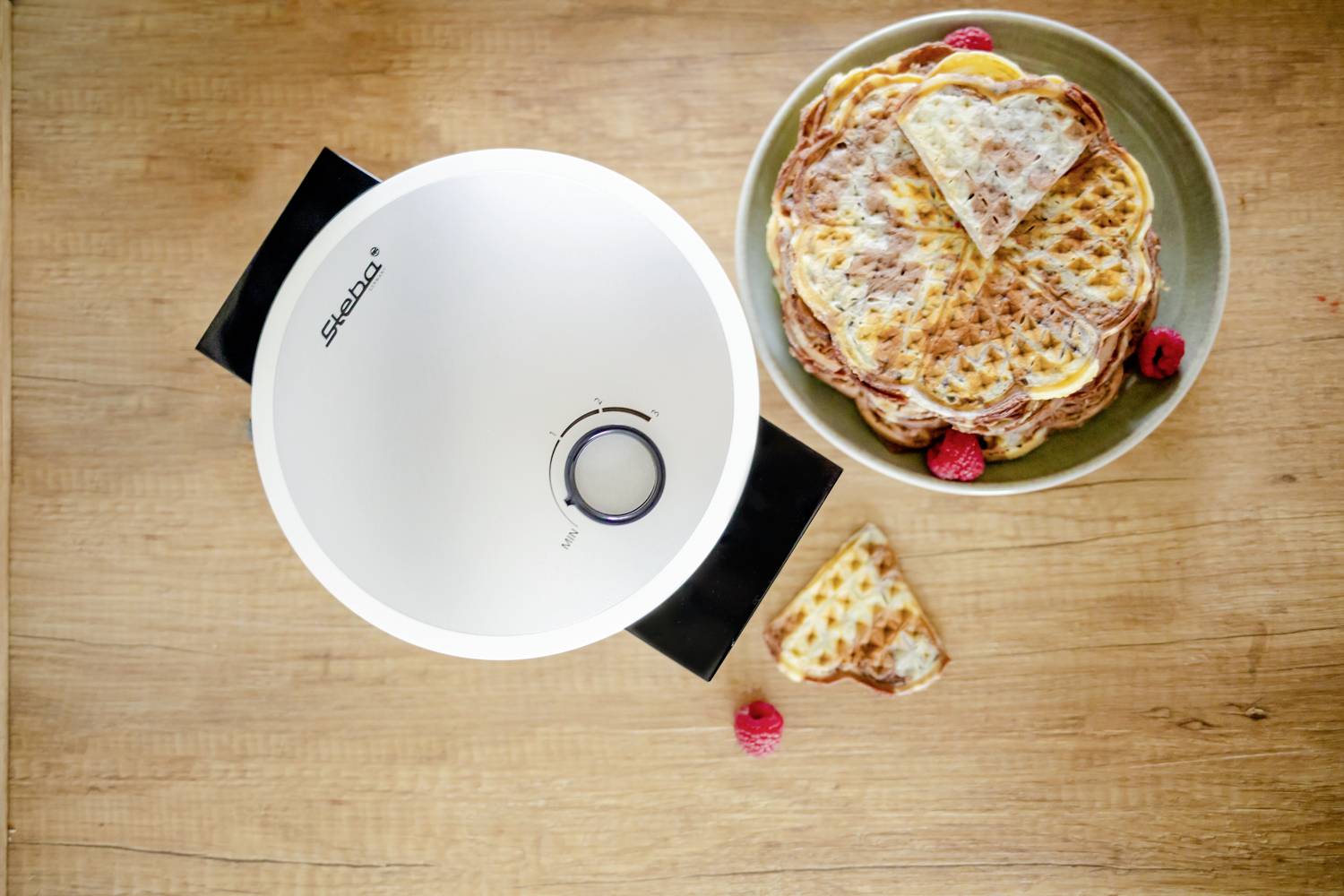 Ein Waffeleisen steht geöffnet neben einem Teller mit gestapelten Waffeln und ein paar Himbeeren auf einem Holztisch.