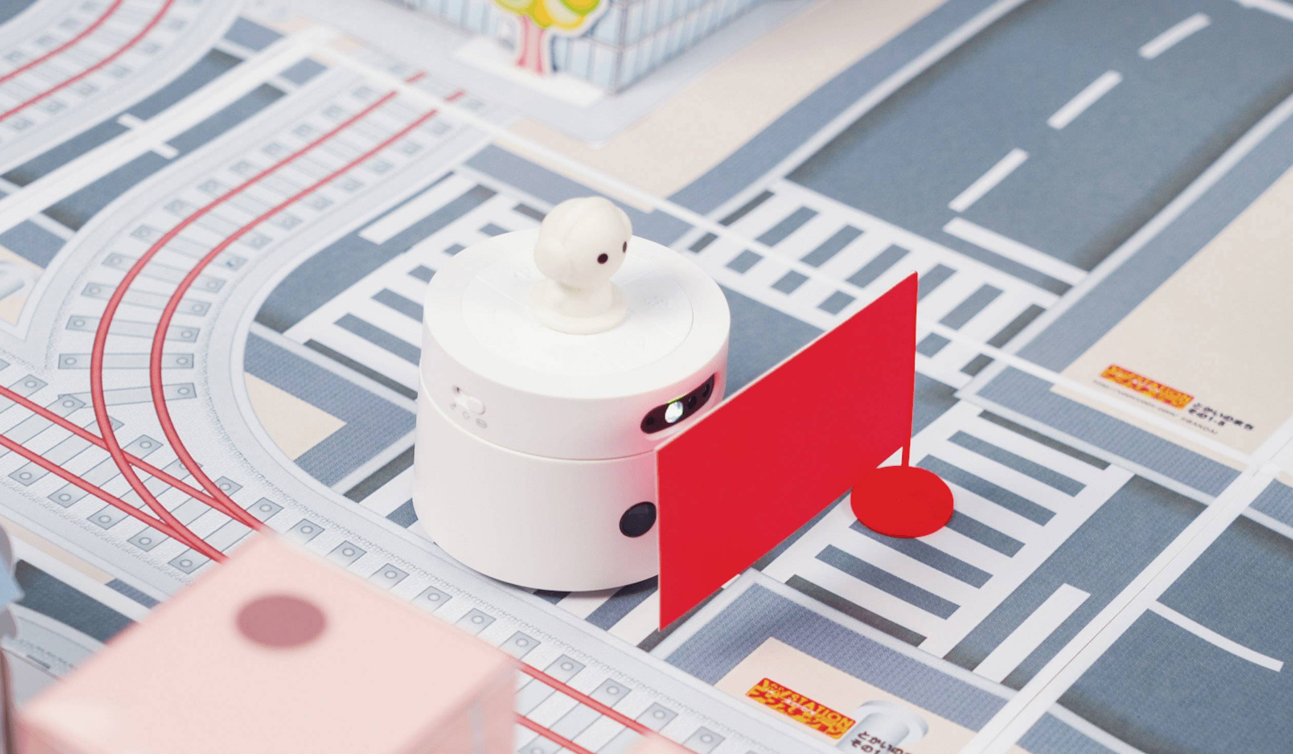 Ein kleiner weißer Roboter navigiert auf einem Spielbrett mit Straßenmotiven, vorbei an einer roten Absperrung.