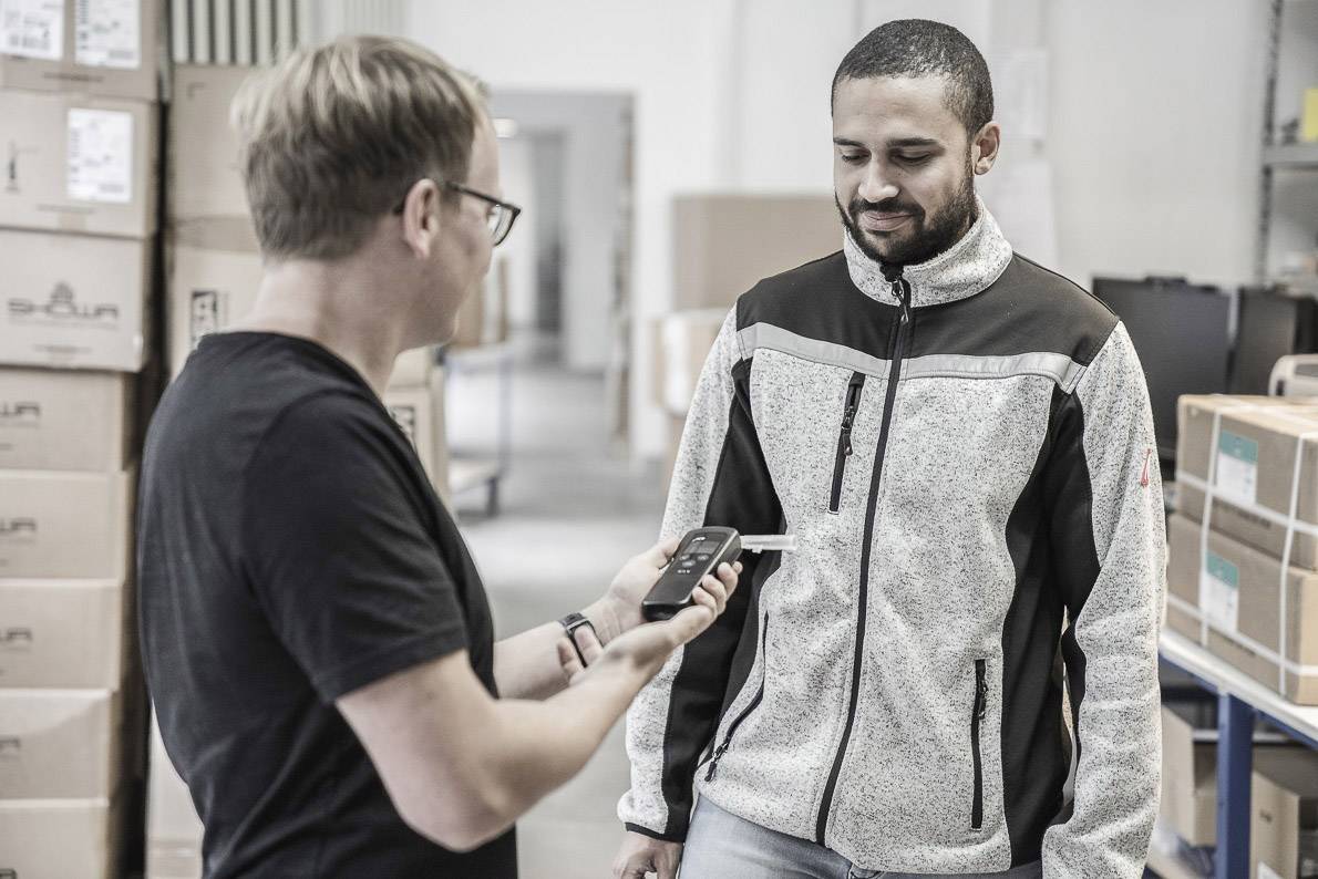 Eine Person scannt den Code eines Pakets in einem Lager, während eine andere Person daneben steht. Im Hintergrund sind Kartons zu sehen.