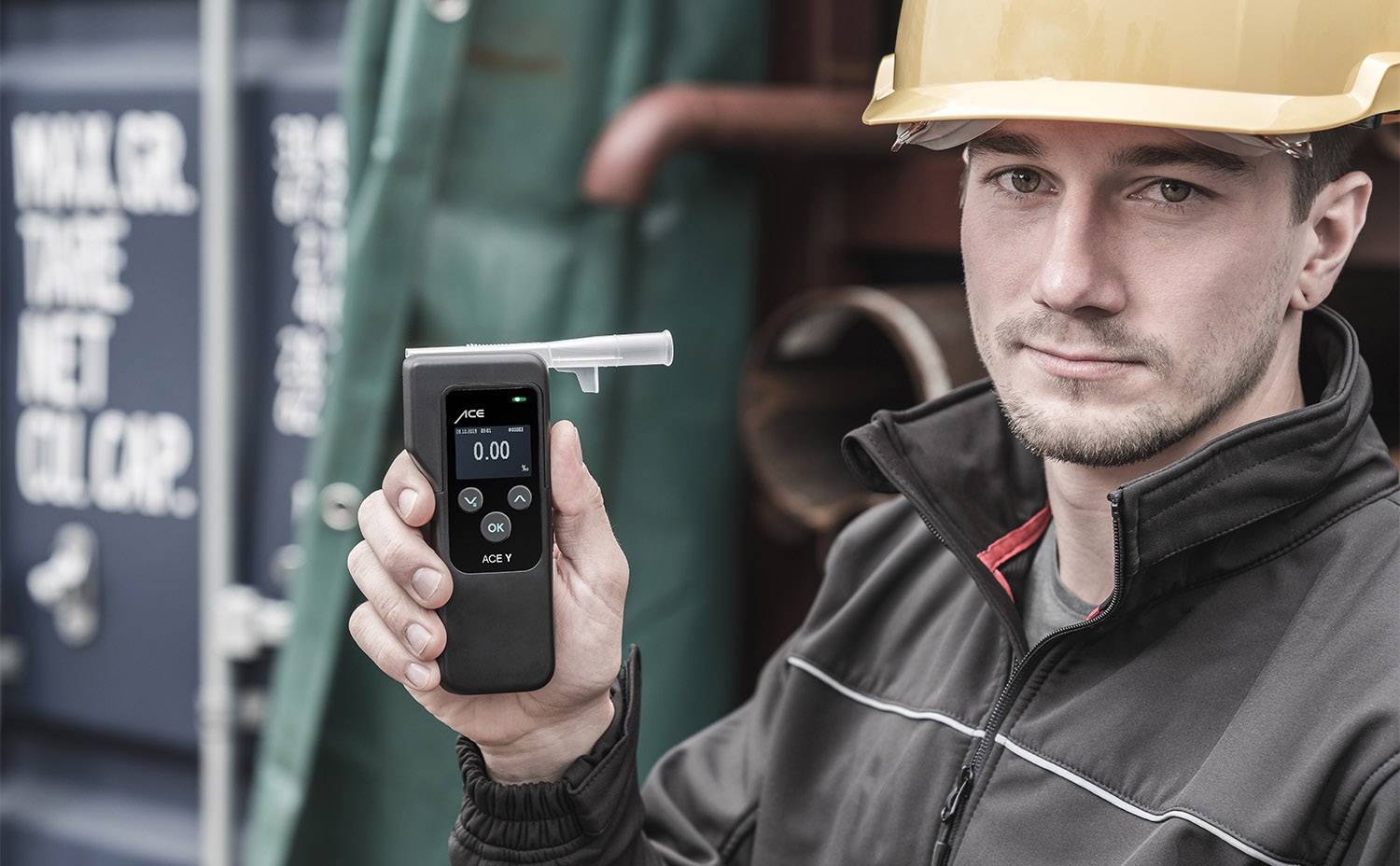 Ein Mann in Arbeitskleidung und Schutzhelm hält ein Alkoholmessgerät in der Hand. Der Hintergrund zeigt einen Container.