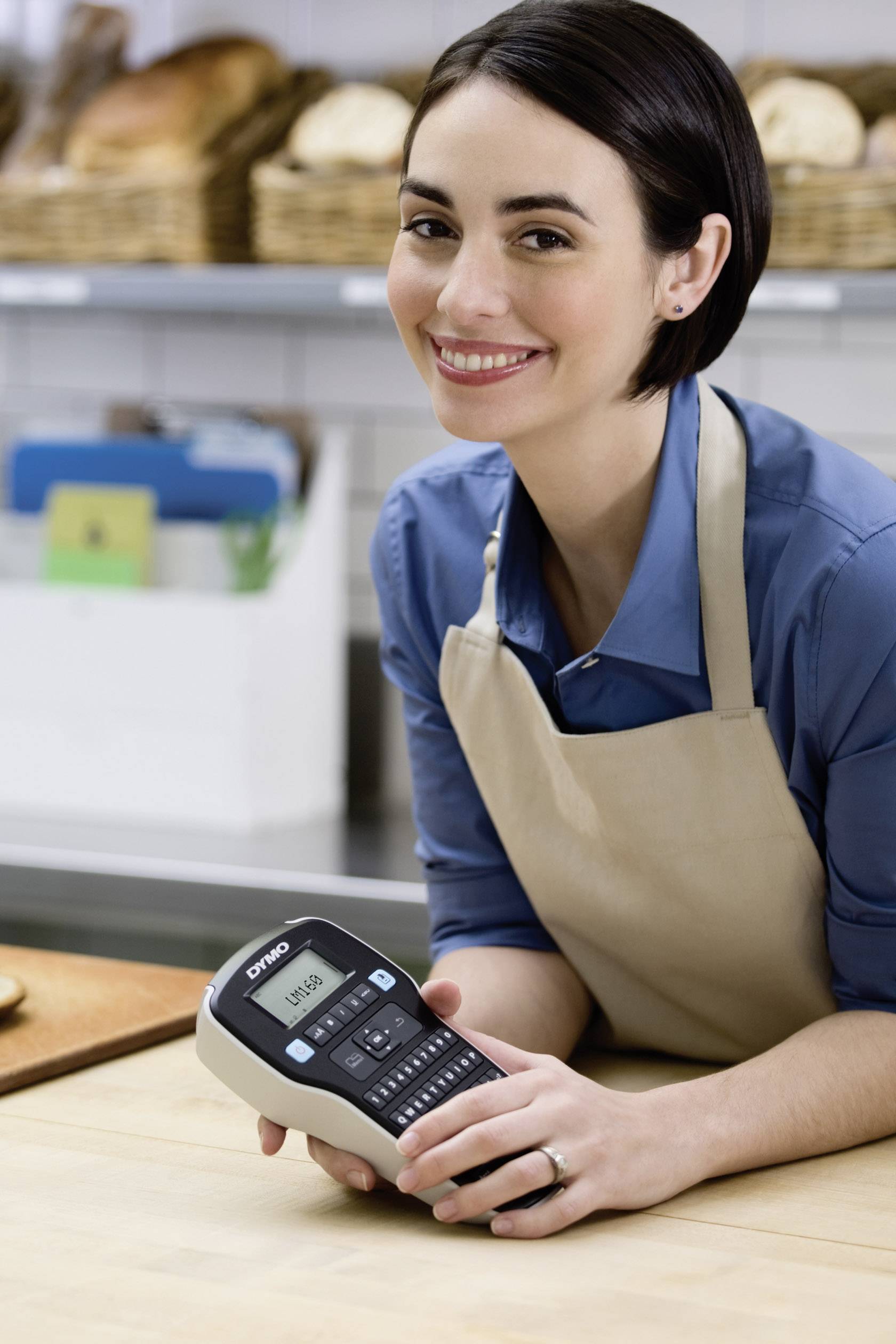 Eine Frau in einer Bäckerei trägt eine Schürze und hält ein Beschriftungsgerät in der Hand. Im Hintergrund sind Brote zu sehen.