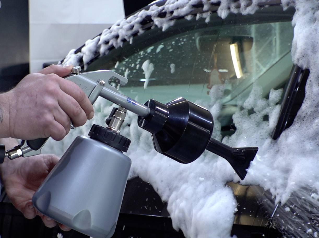 Eine Person verwendet einen Schaumsprüher, um eine Autoscheibe zu reinigen und dicken seifigen Schaum aufzutragen.