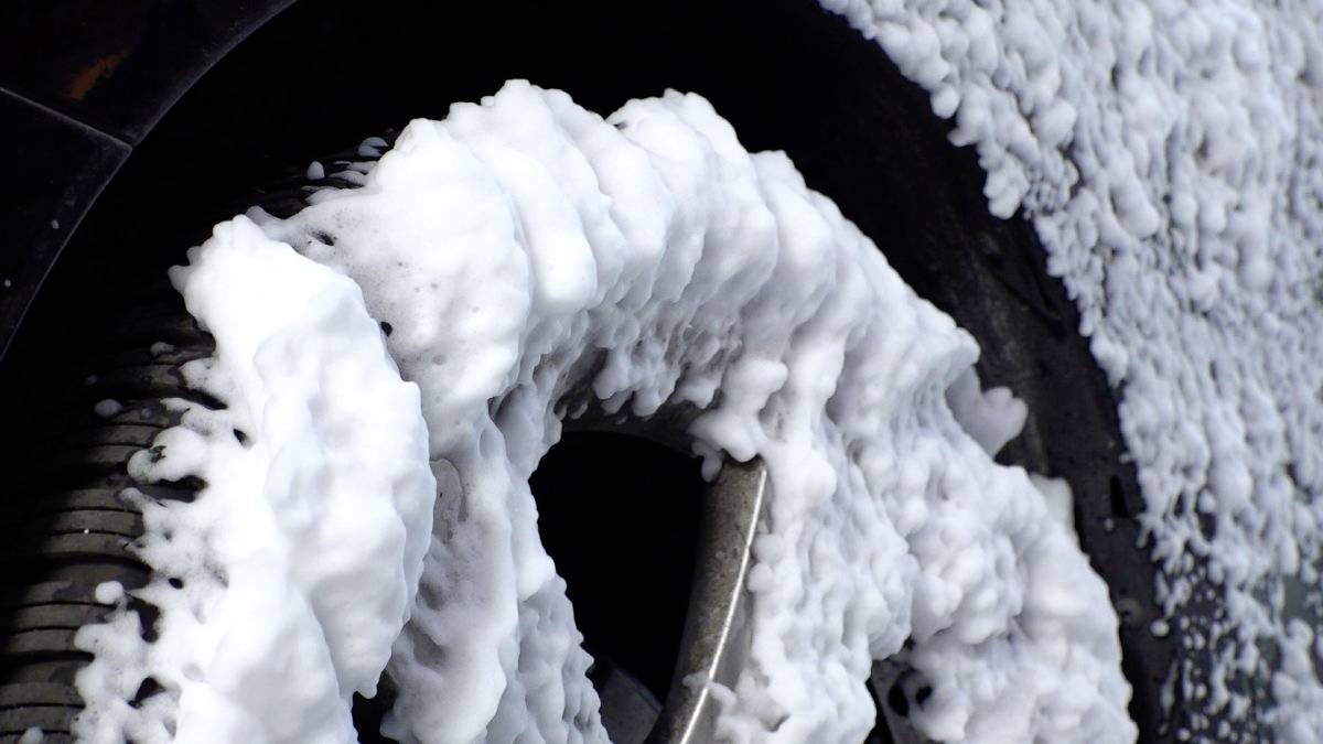 Nahaufnahme eines Autorads, das während eines Reinigungsprozesses mit dichtem weißem Seifenschaum bedeckt ist. Der Schaum haftet am Reifen und teilweise an der Felge.