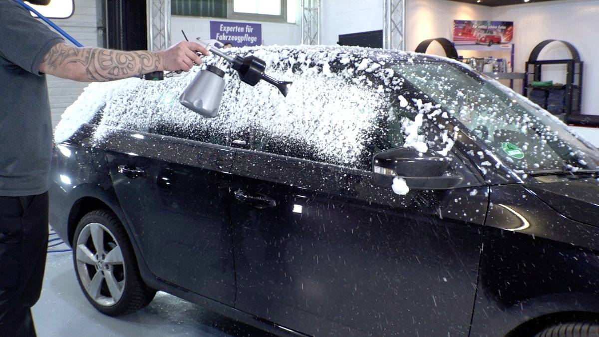 Eine Person trägt mit einer Spritzpistole Schaum auf ein schwarzes Auto in einer Autoaufbereitungswerkstatt auf. Der Fokus liegt auf der Autoseite, die größtenteils mit weißem Schaum bedeckt ist.