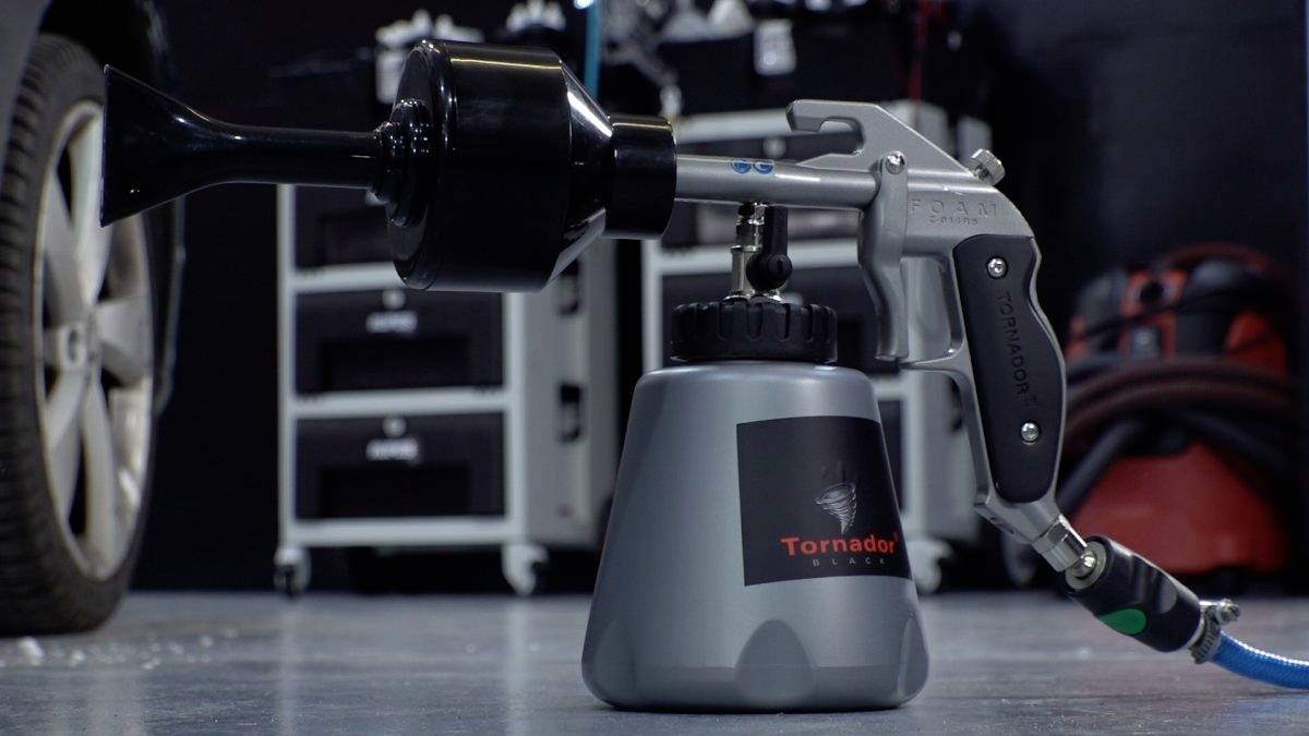Ein Autoaufbereitungswerkzeug mit einer Düse und einem Behälter, der mit 'Tornador' beschriftet ist, steht auf dem Garagenboden in der Nähe eines Autoreifens und weiterer Ausrüstung im Hintergrund.