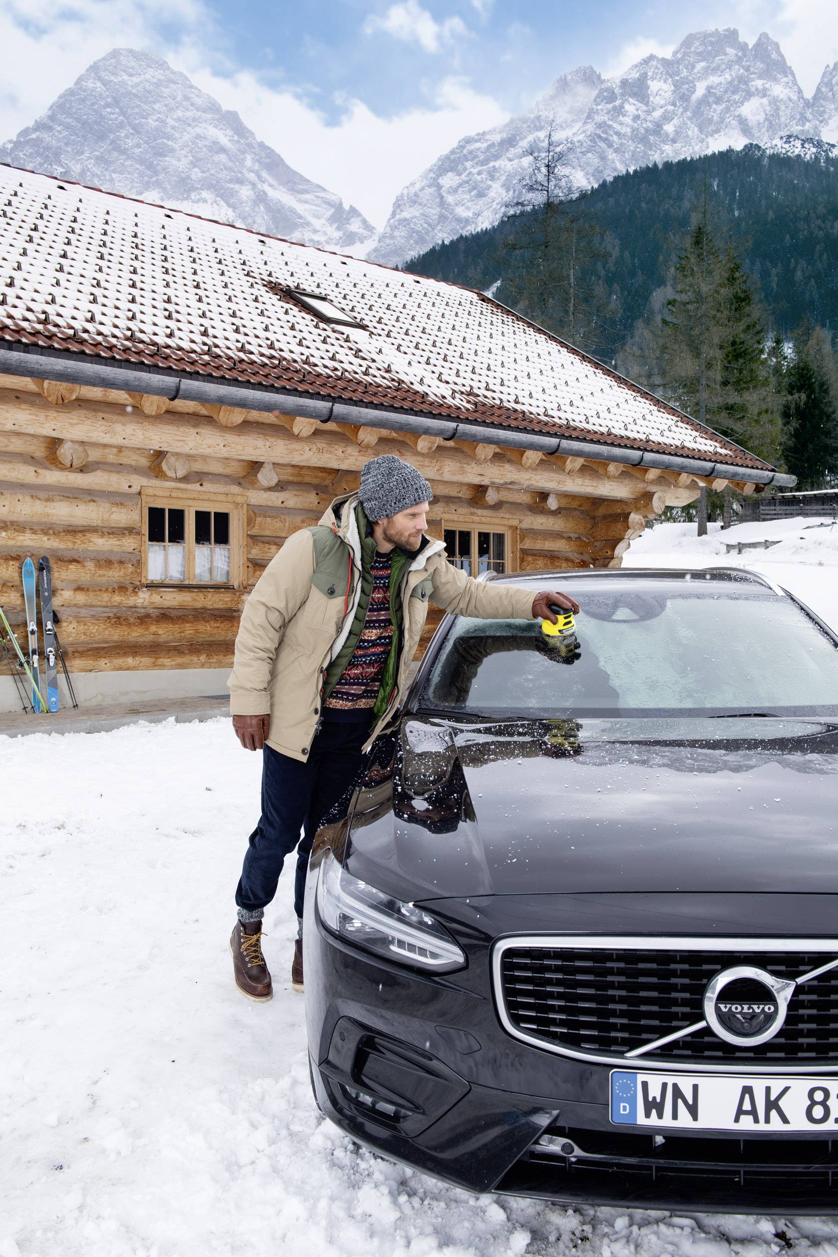 Ein Mann kratzt im Schnee vor einer Holzhütte und Bergen Eis von der Windschutzscheibe eines grauen Autos ab. Skier lehnen an der Hütte.