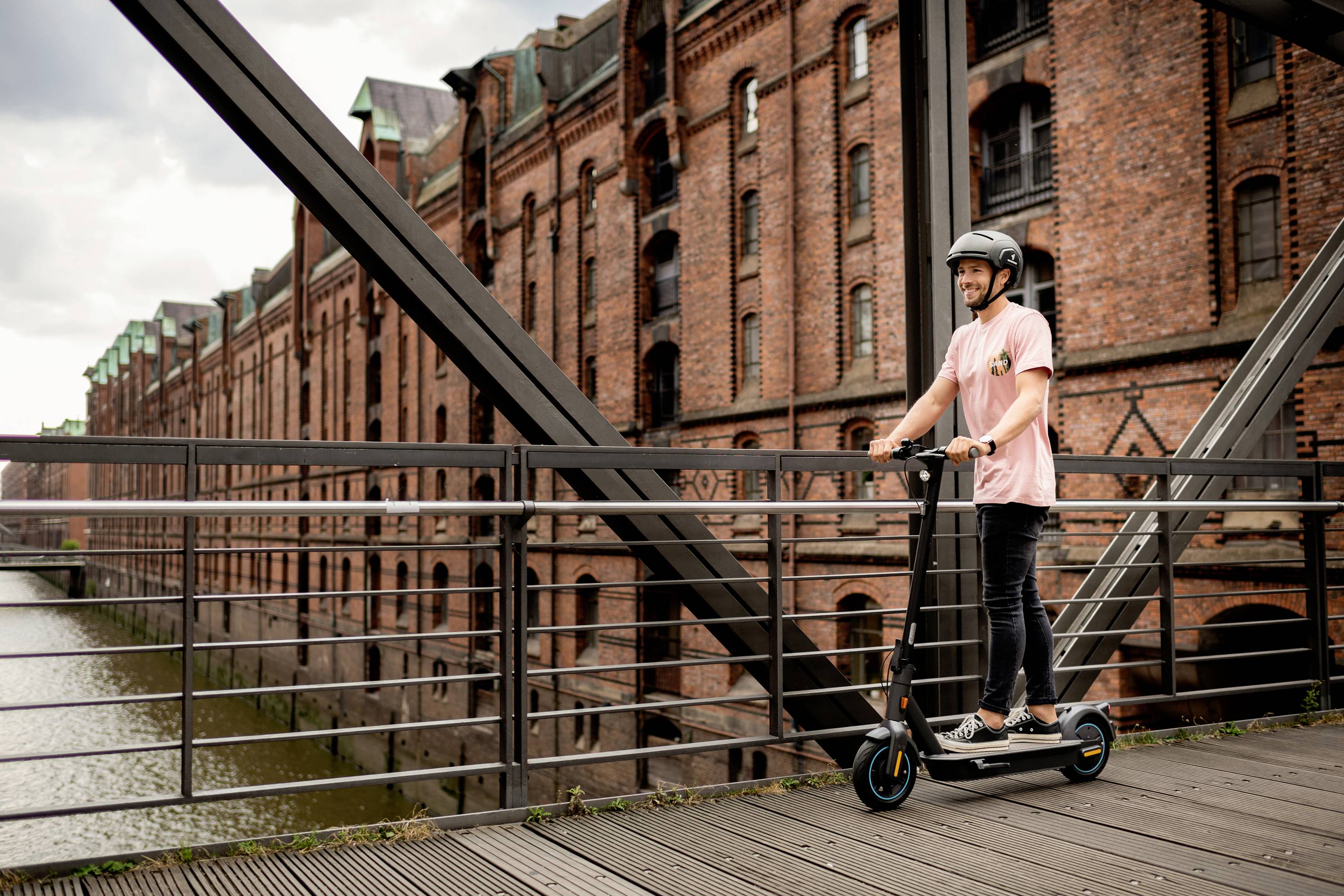 Eine Person fährt mit einem Elektroroller über eine Brücke vor einem alten Backsteingebäude.