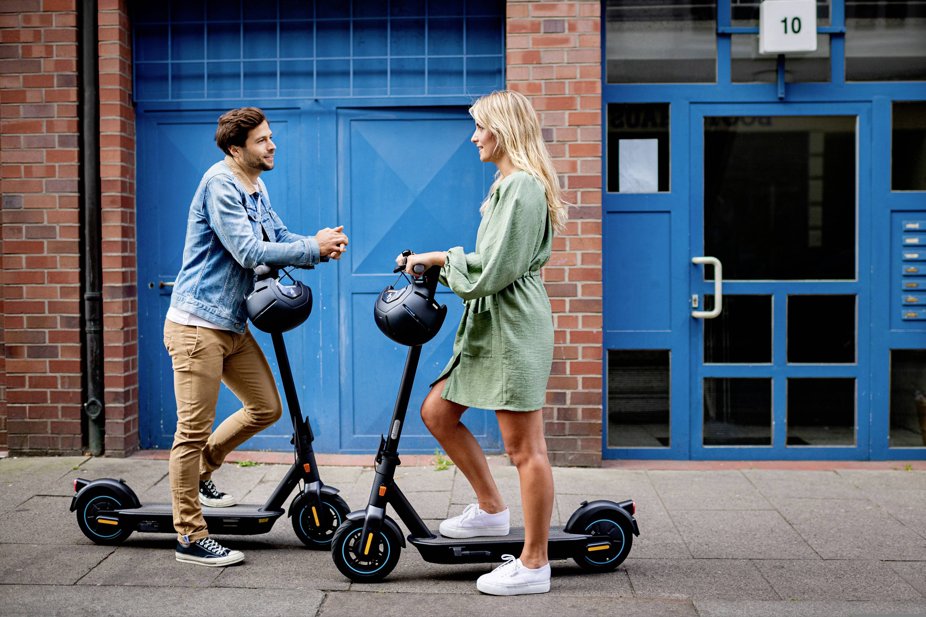 Ein Mann und eine Frau stehen sich gegenüber, lehnen sich auf E-Scooter. Sie unterhalten sich vor einem Gebäude mit blauen Türen.