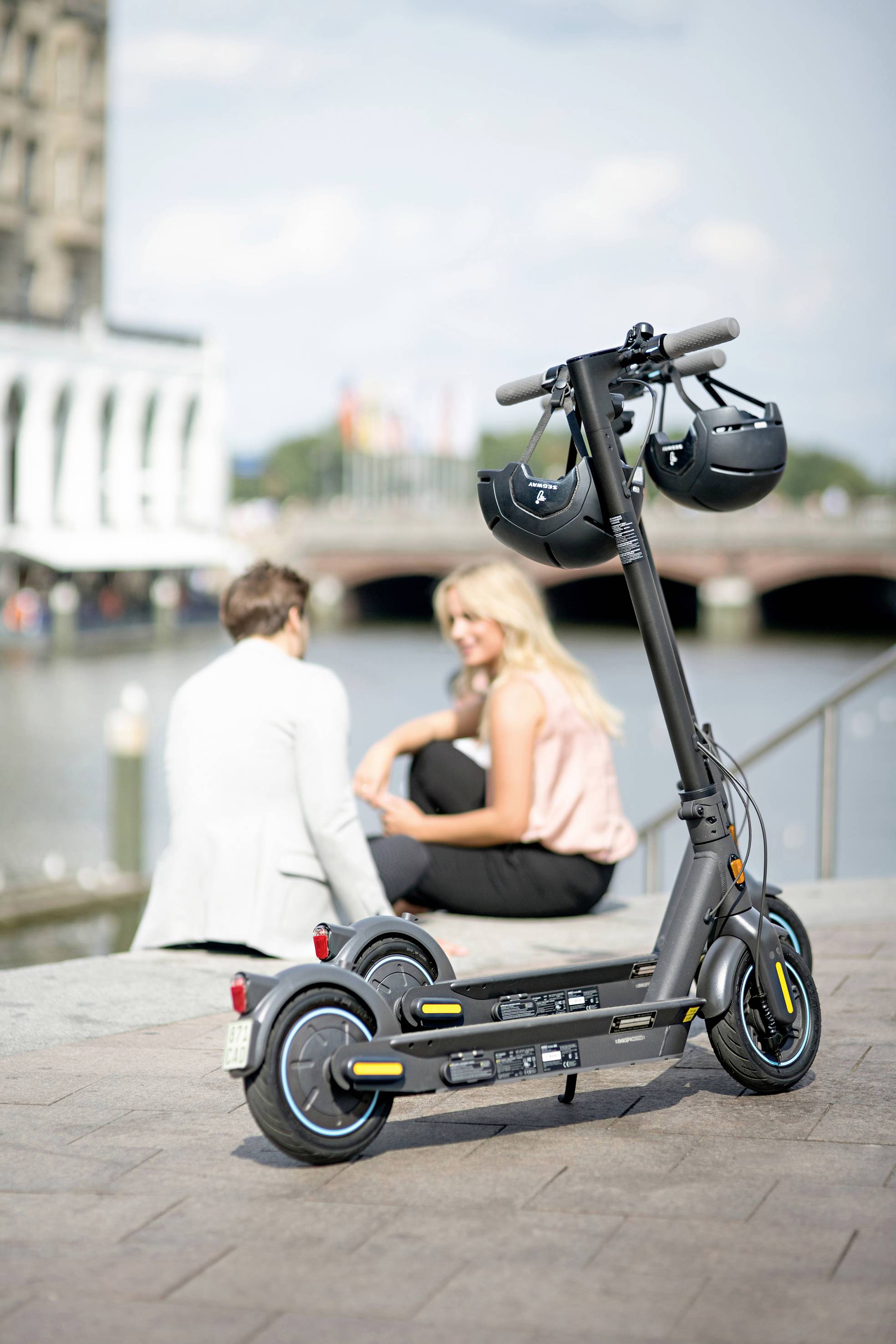 Ein schwarzer E-Scooter mit zwei Helmen steht im Vordergrund an einem Fluss. Im Hintergrund sitzen zwei Personen auf einer Mauer.