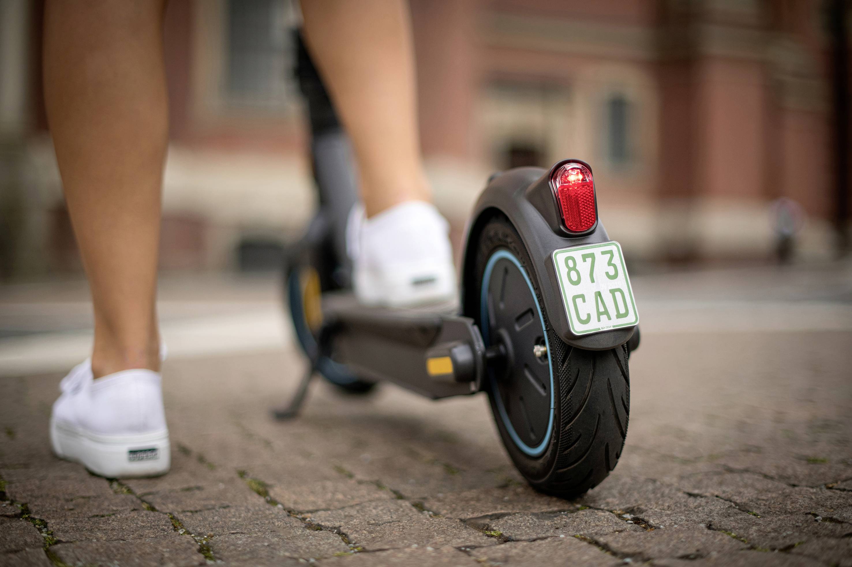 Eine Person fährt auf einem E-Scooter über einen gepflasterten Gehweg. Der Fokus liegt auf dem Hinterrad und dem Nummernschild '873 CAD'.