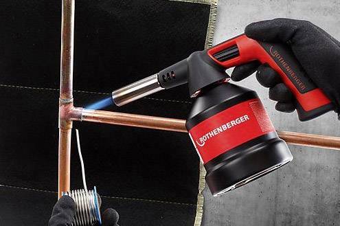 'Eine Lötpistole erhitzt ein Kupferrohr. Eine Person hält Lötzinn nahe an die erhitzte Stelle, um ein Verbindungsstück zu löten.'