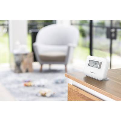 Ein digitales Raumthermostat zeigt 21 Grad Celsius an und steht auf einer Holzoberfläche. Im Hintergrund verschwommene Möbel.