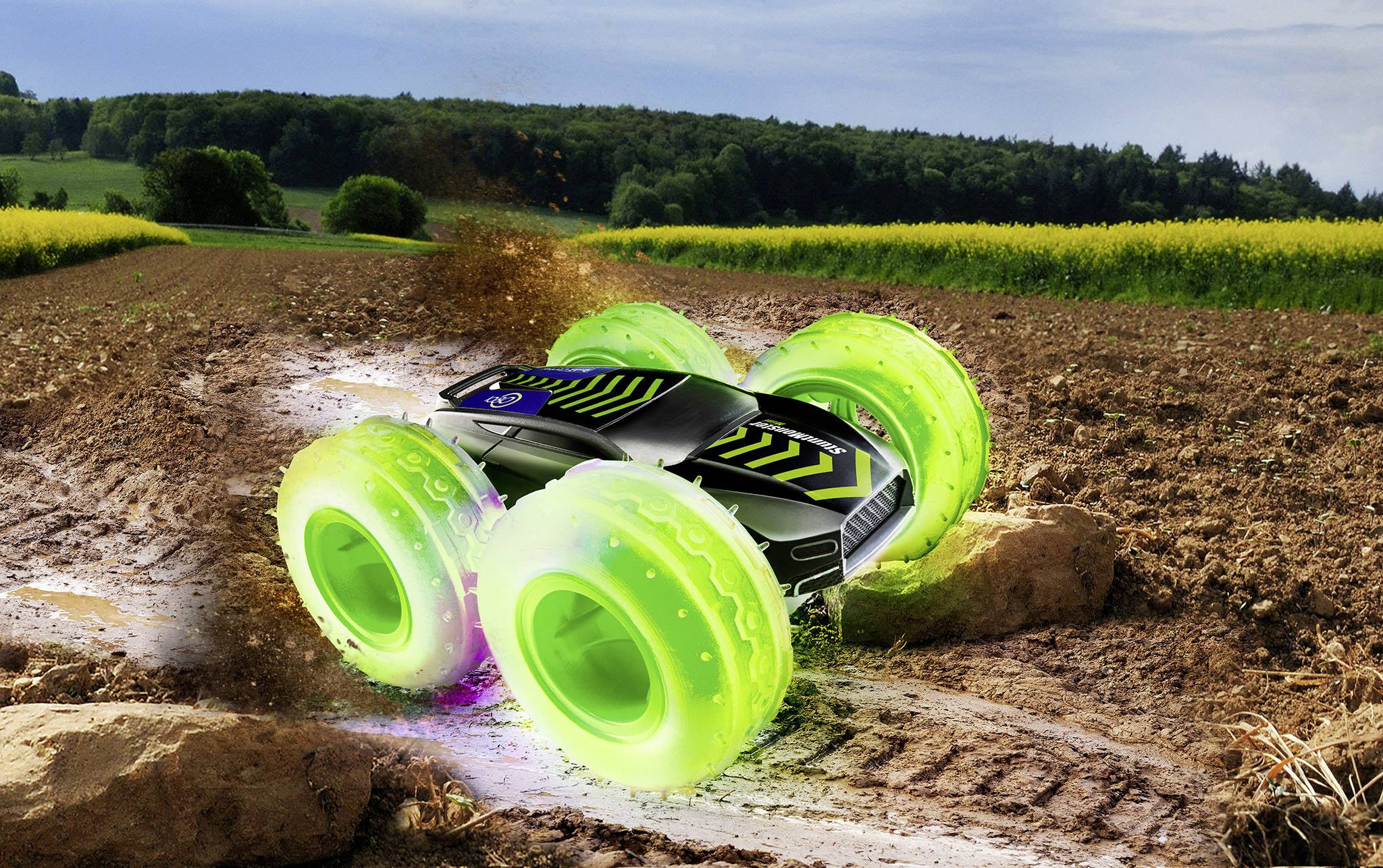 Spielzeugauto mit großen, leuchtend grünen Rädern fährt durch schlammiges Feld, wirbelt Erde und Wasser auf. Hintergrund: Wald und Felder.