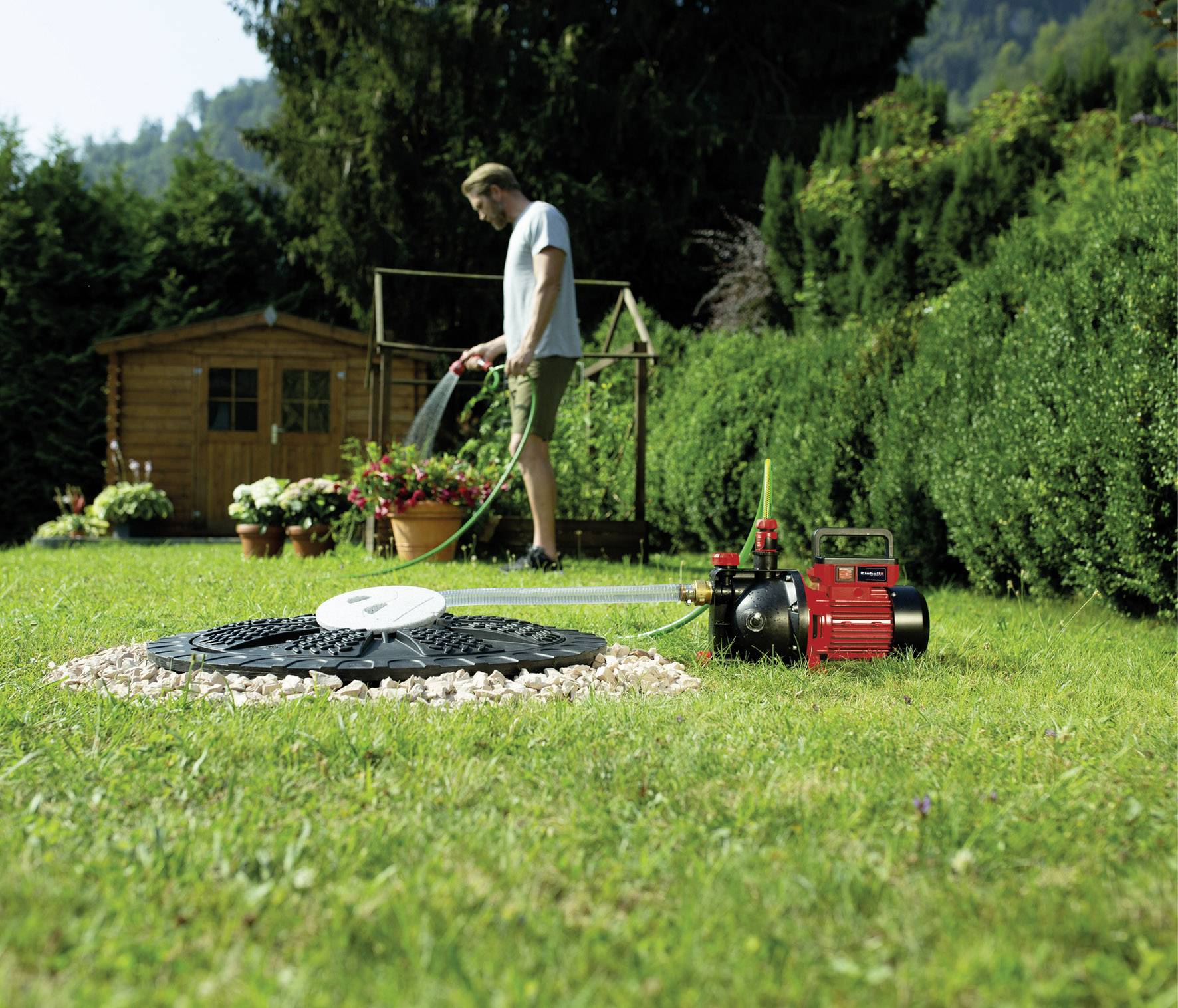 Ein Mann gießt mit einem Schlauch Pflanzen in einem Garten; im Vordergrund ein schwarzer Gartensprenger und eine rote Pumpe.