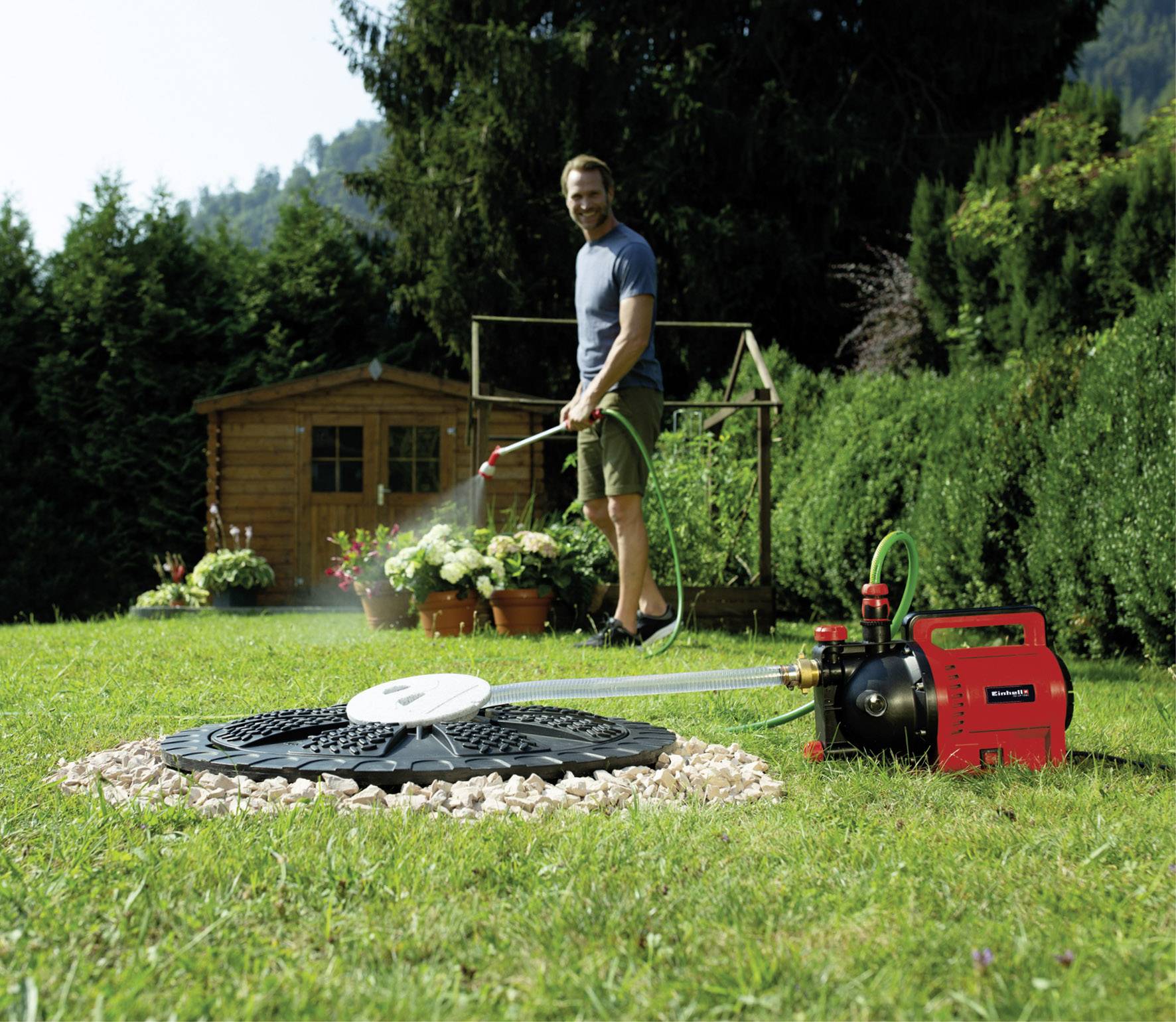 Ein Mann im Garten gießt Pflanzen mit einem Schlauch. Eine rote Wasserpumpe ist im Vordergrund sichtbar. Hintergrund: Gartenhaus, Bäume.