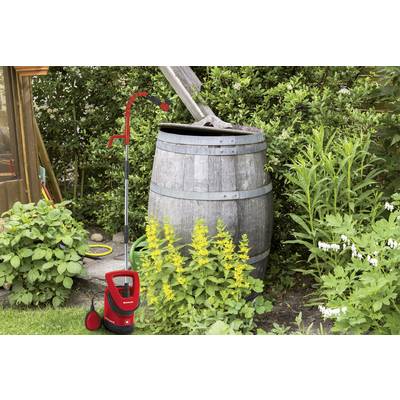 Ein alter Holzfass mit einem offenen Deckel steht in einem Garten, umgeben von grünen Pflanzen und einem roten Wasserpumpengerät daneben.