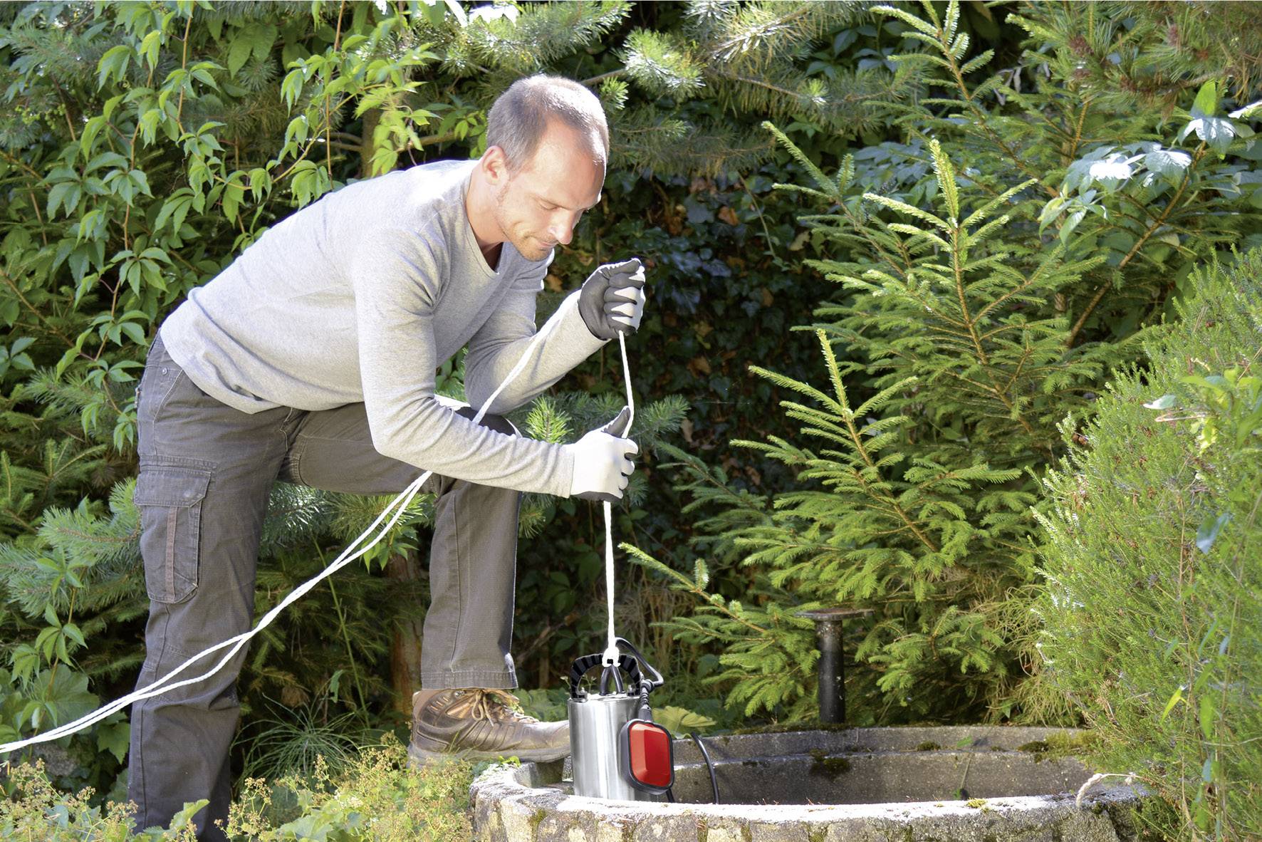 Eine Person hantiert mit einem elektrischen Gerät an einem Brunnen in einem Garten, umgeben von Bäumen und Sträuchern.