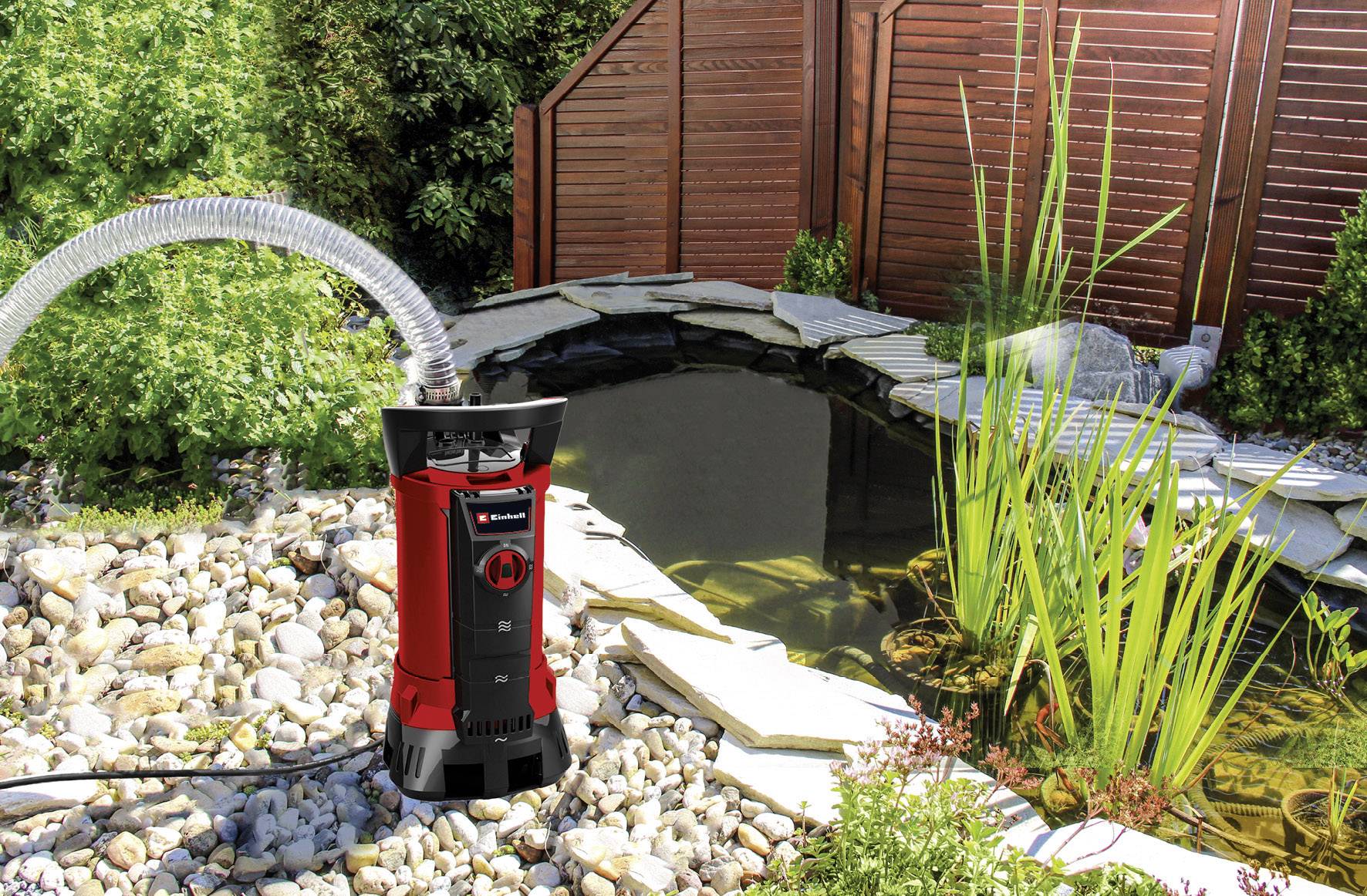 Ein Teich im Garten mit Umwälzpumpe links, umgeben von Steinen und Pflanzen, Holzzaun im Hintergrund.
