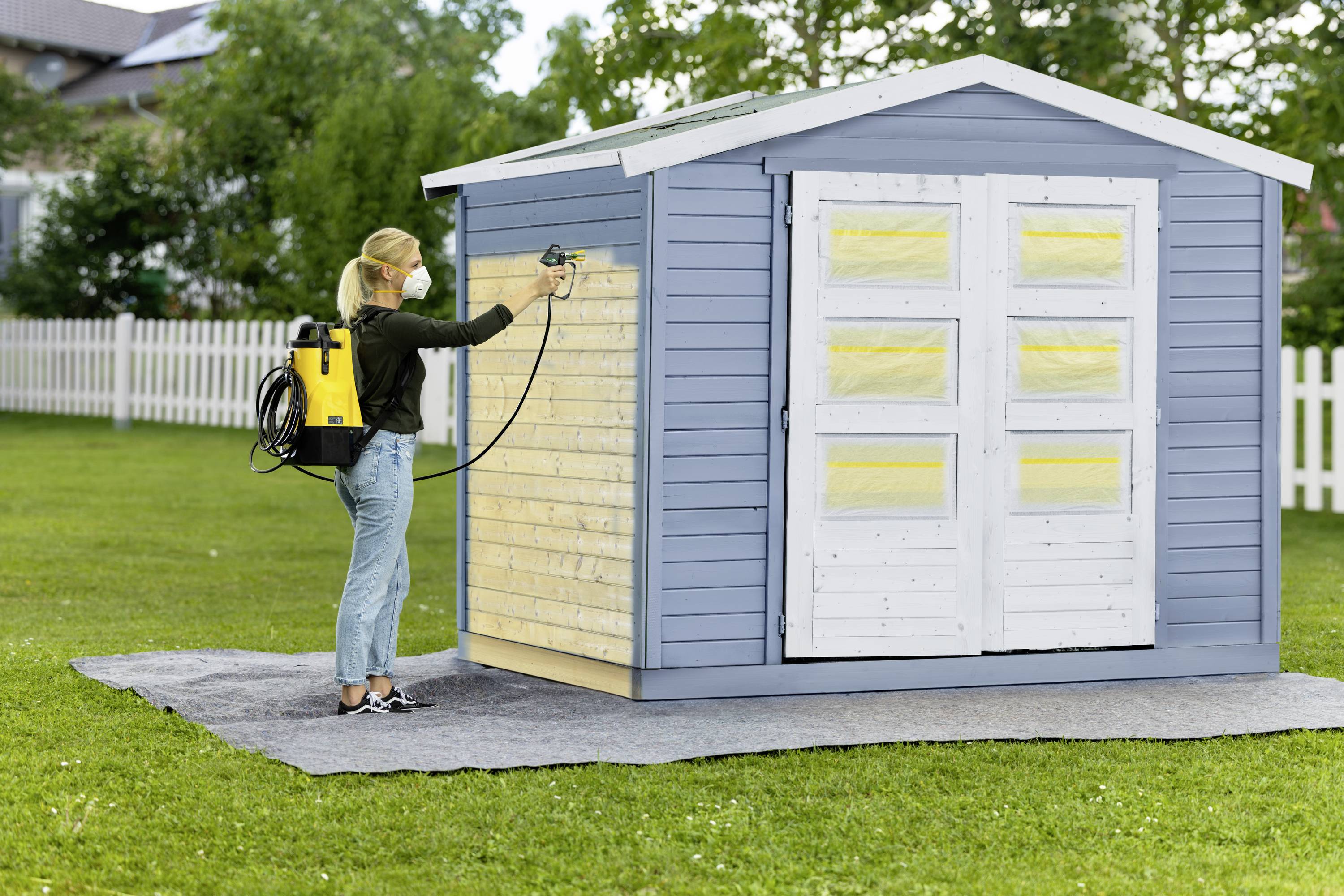 Eine Person sprüht Farbe auf die Holzwand eines blauen Gartenhauses im Freien. Sie trägt einen Farbsprüher auf dem Rücken.
