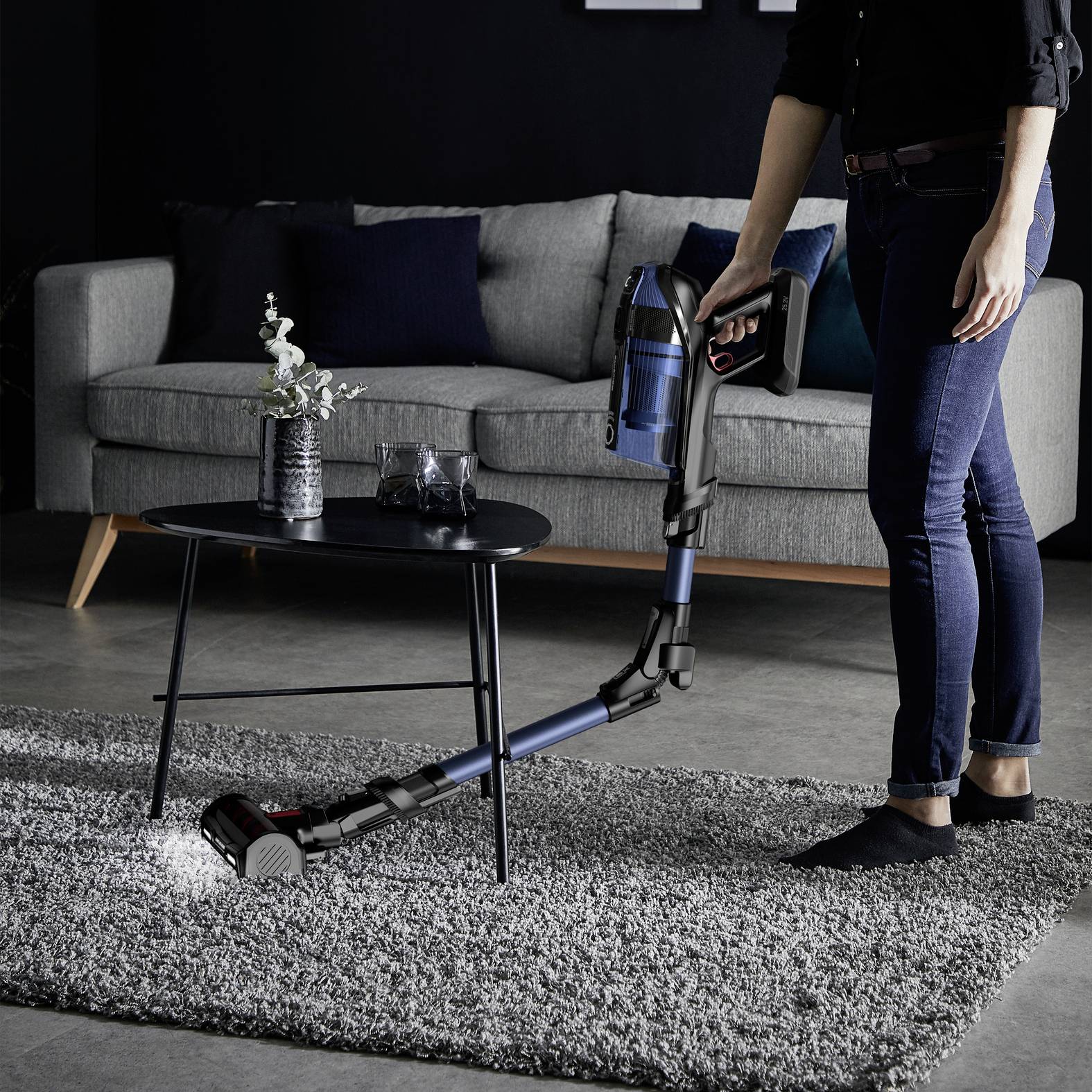 Eine Person benutzt einen kabellosen Staubsauger auf einem Teppich in einem modernen Wohnzimmer mit einem Sofa und einem Couchtisch.