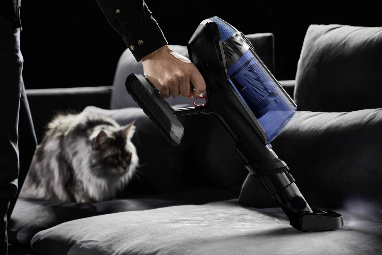 Eine Person saugt mit einem Handstaubsauger Tierhaare von einem grauen Sofa, während eine Katze aufmerksam zuschaut.