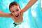 Ein lachender Junge mit nassen Haaren schwimmt im Pool und hebt freudig die Arme.