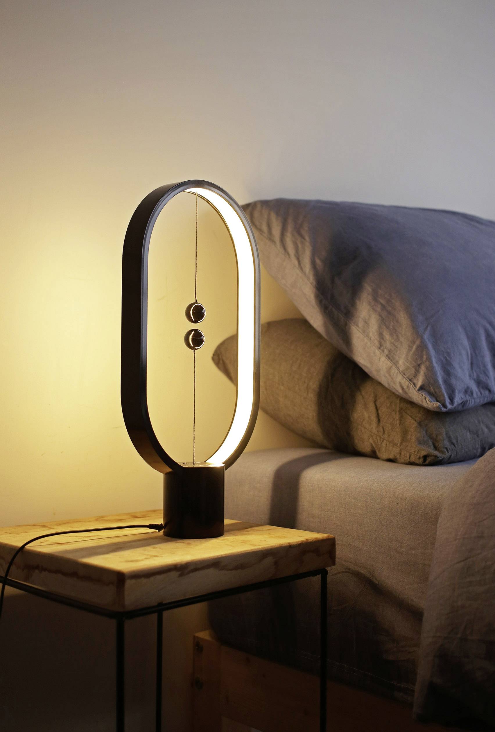 Eine moderne Tischlampe mit ovalem Rahmen und Schwebeschalter steht auf einem Nachttisch neben einem Bett mit grauer Bettwäsche.
