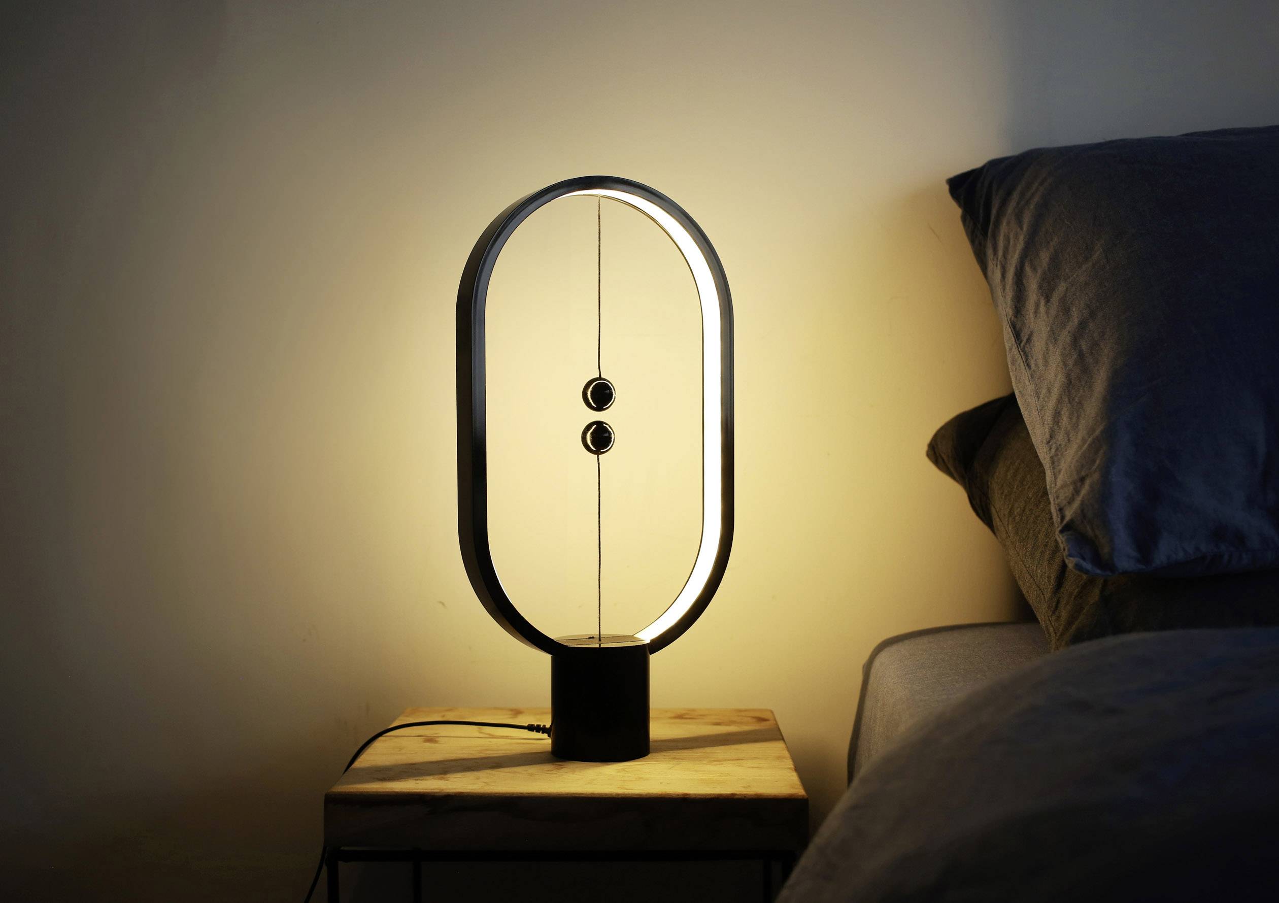 Eine moderne Tischlampe mit ovalem Rahmen und zwei schwebenden Kugeln als Schalter steht auf einem Nachttisch neben einem Bett.