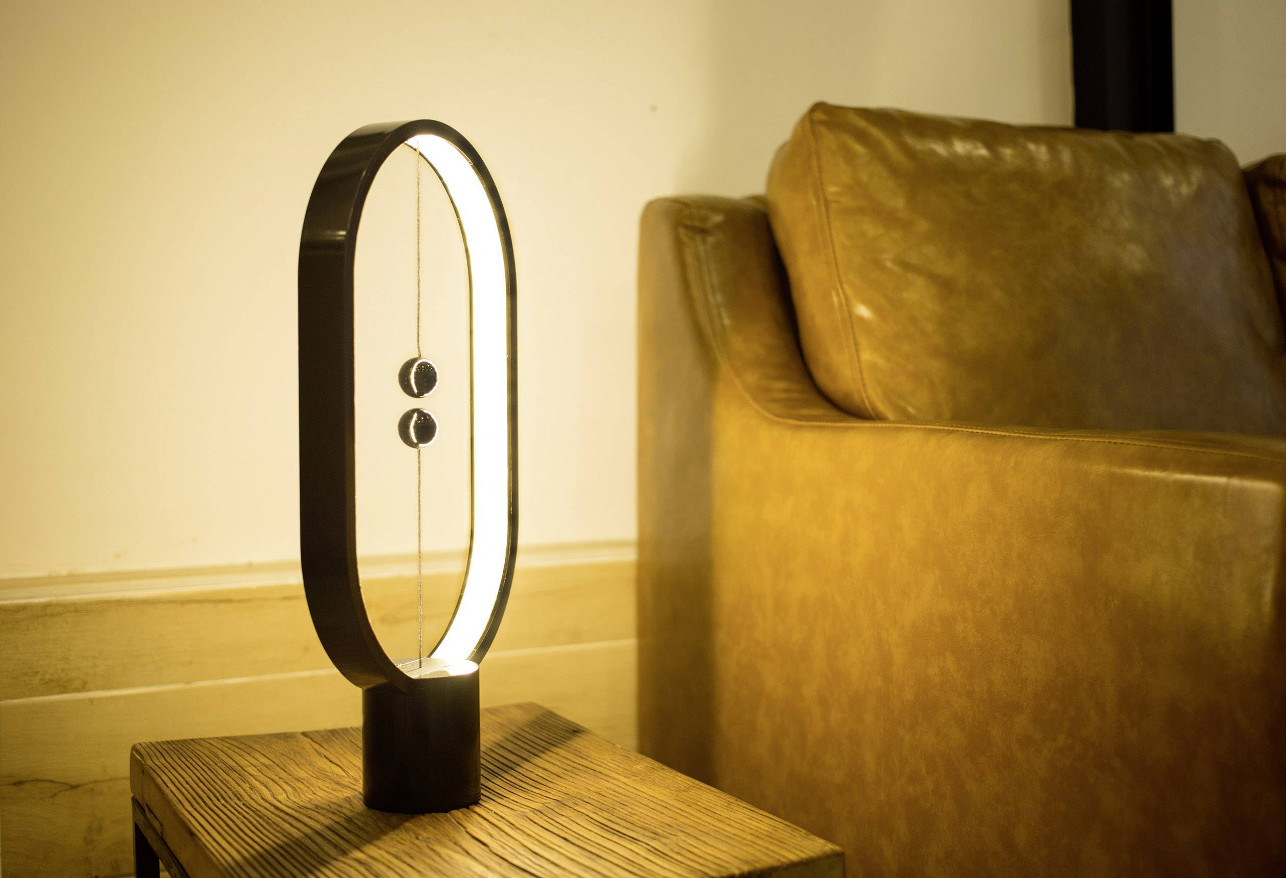 Moderne ovale Tischlampe auf Holzfläche, weiches Licht spendend. Im Hintergrund ein brauner Ledersessel an einer Wand.